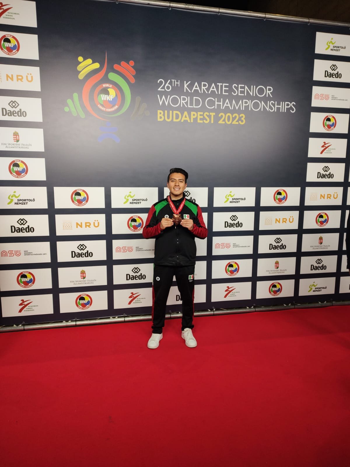 Persona en un evento de karate, sosteniendo una medalla, con un fondo que dice "26th Karate Senior World Championships, Budapest 2023".