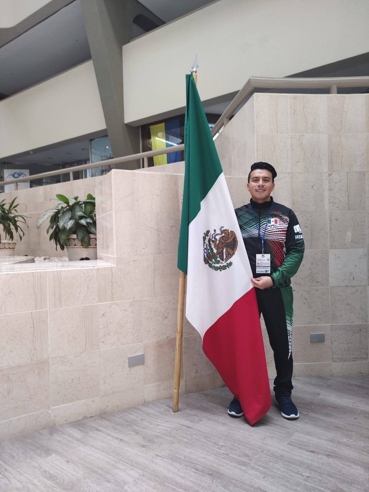 Persona junto a una bandera de México, en un edificio con plantas y diseño interior moderno.