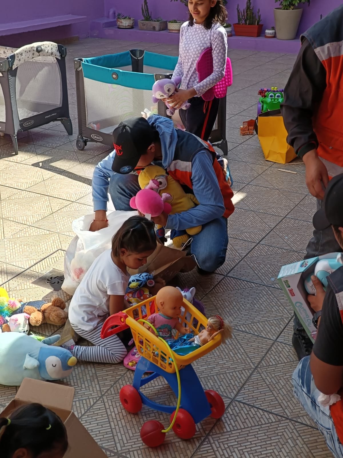 Un grupo de niños y adultos en una habitación decorada con colores brillantes. Los niños están jugando con juguetes, incluidas muñecas, peluches y un carrito de juguete. Hay corrales de bebés y cajas en el suelo. Una niña sostiene un peluche morado,