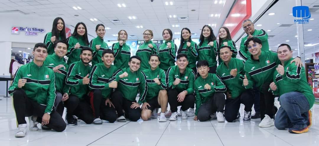 Grupo de personas en un aeropuerto, vestidas con chaquetas verdes, posando para una foto juntos. Al fondo se ven tiendas y luces del techo.