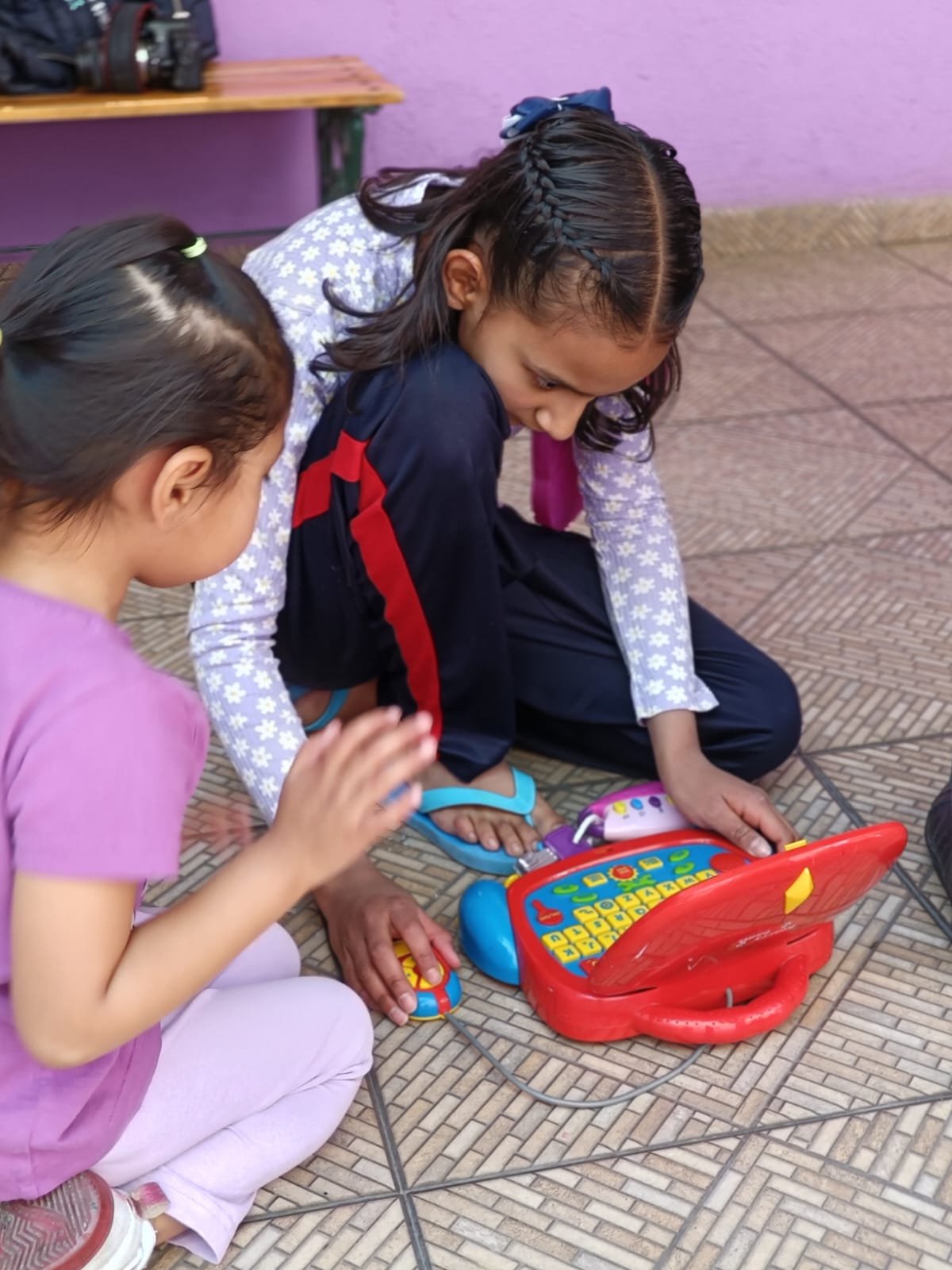 Dos niños jugando con una computadora de juguete en el suelo, en un ambiente interior con paredes y piso de color claro.