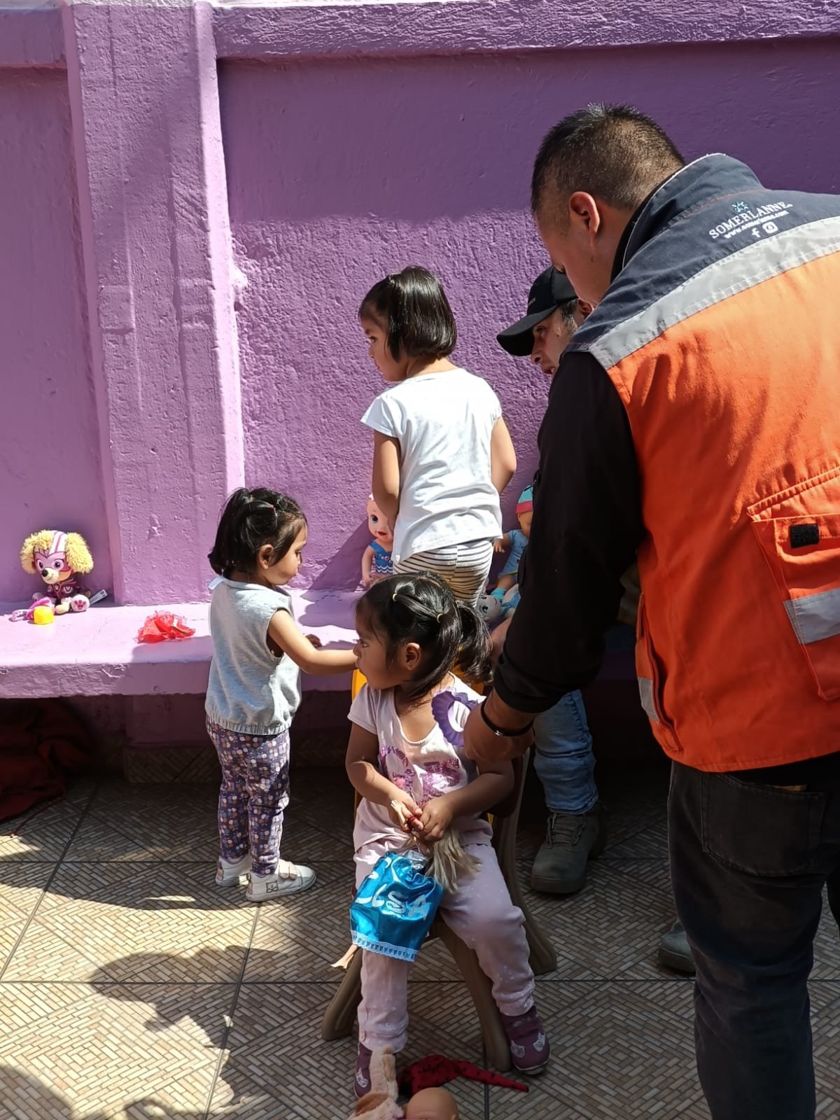 Niños jugando con juguetes en un patio con paredes moradas. Un adulto con chaqueta naranja observando la escena.