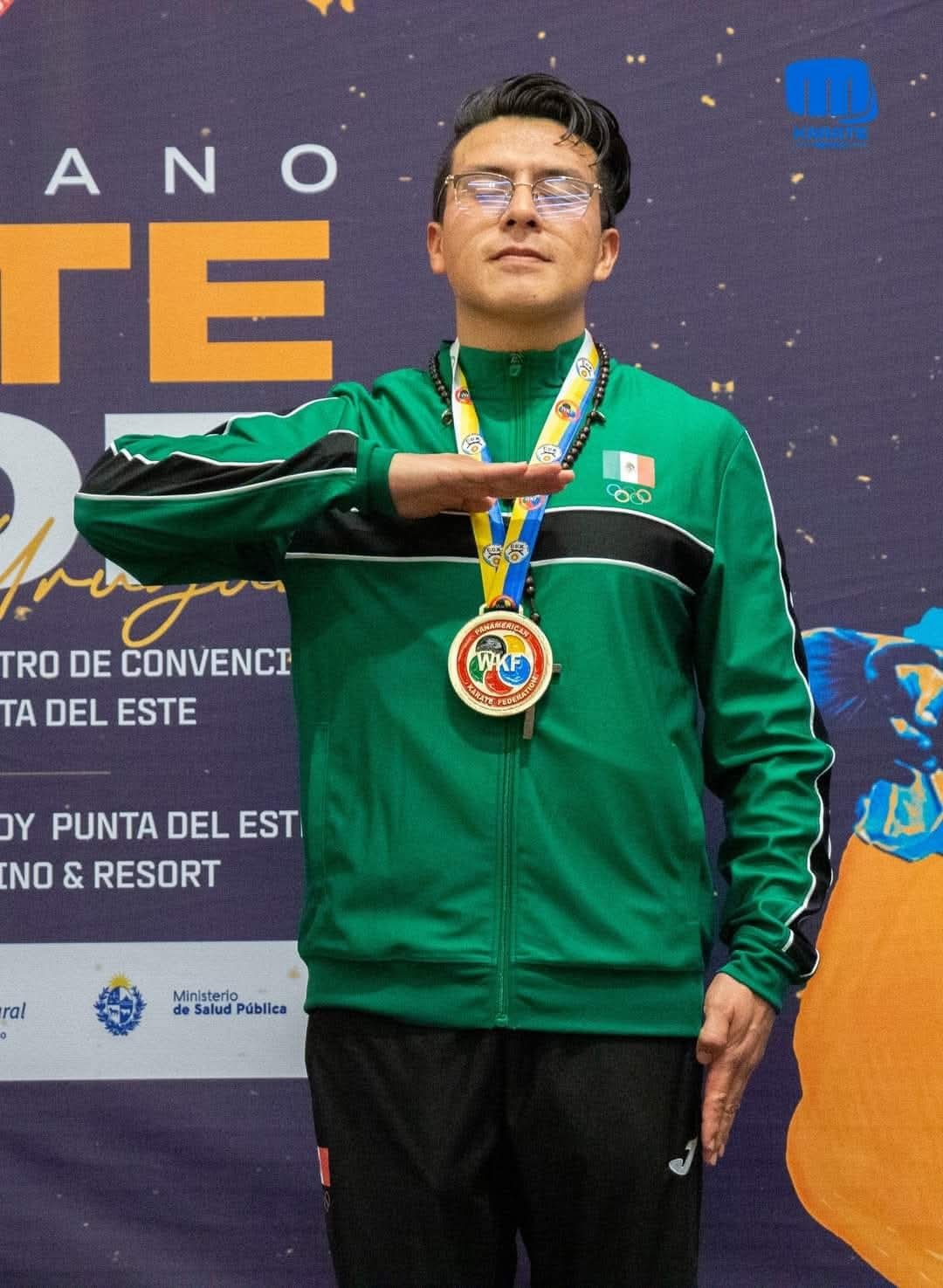 Persona con medalla en ceremonia de premiación, usando chaqueta verde con bandera y símbolos olímpicos, fondo con logotipos y texto.