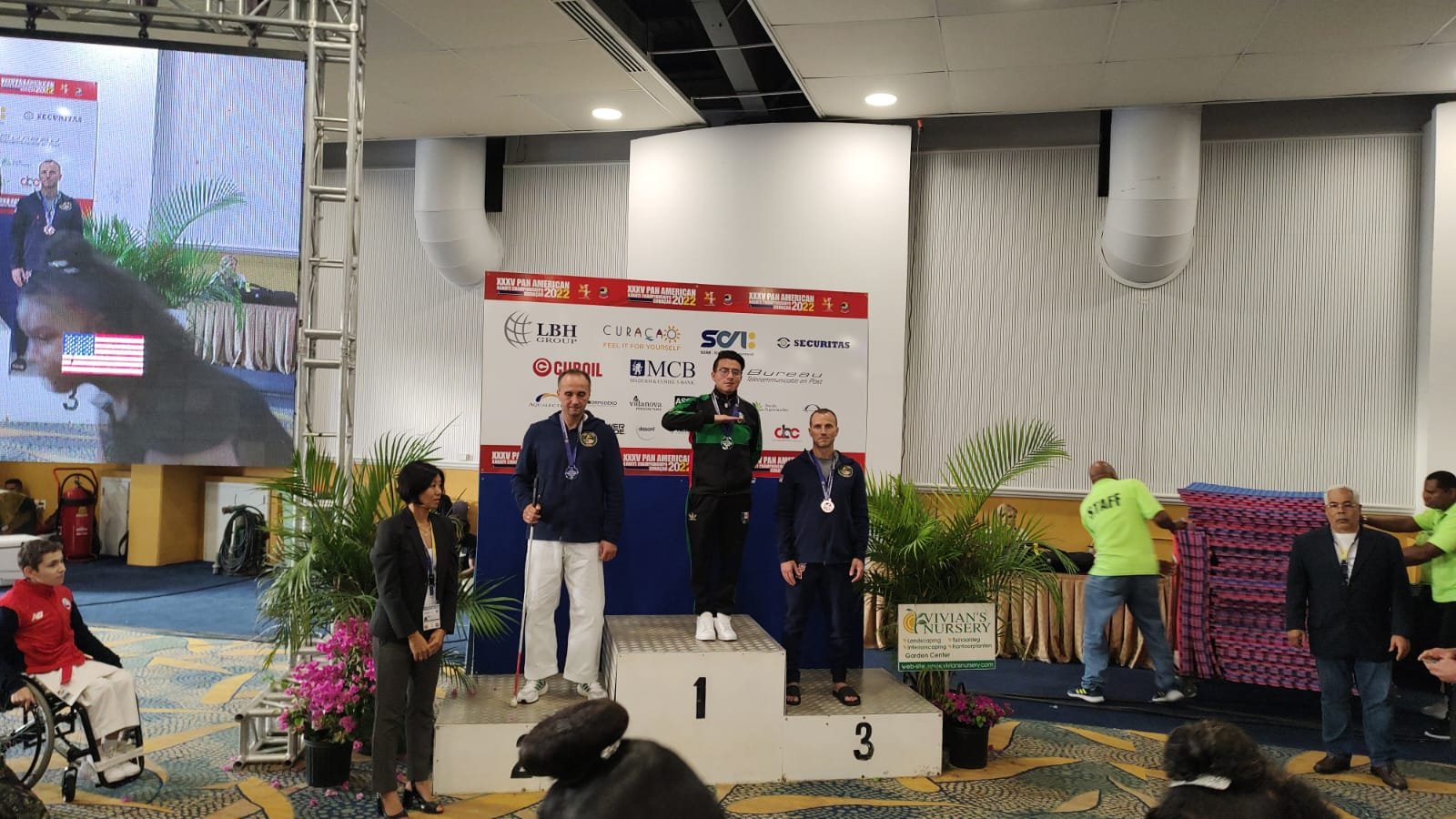 Ceremonia de entrega de medallas con tres hombres en un podio. Un hombre en ropa deportiva verde oscuro está en el centro en el primer lugar. A su lado, dos hombres en sudaderas azules presentan medallas alrededor de sus cuellos, ocupando el segundo