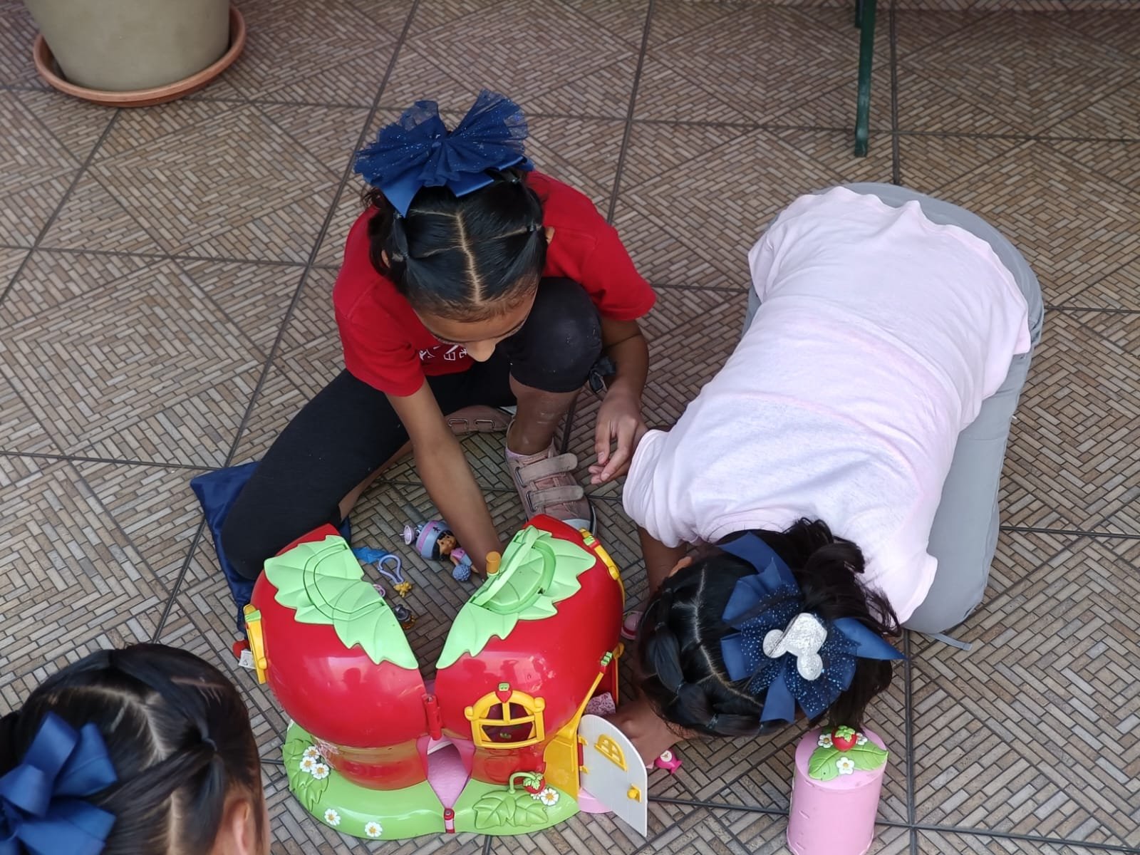 Niños jugando con una casa de juguete en forma de fresas grandes, sobre un piso de azulejos.