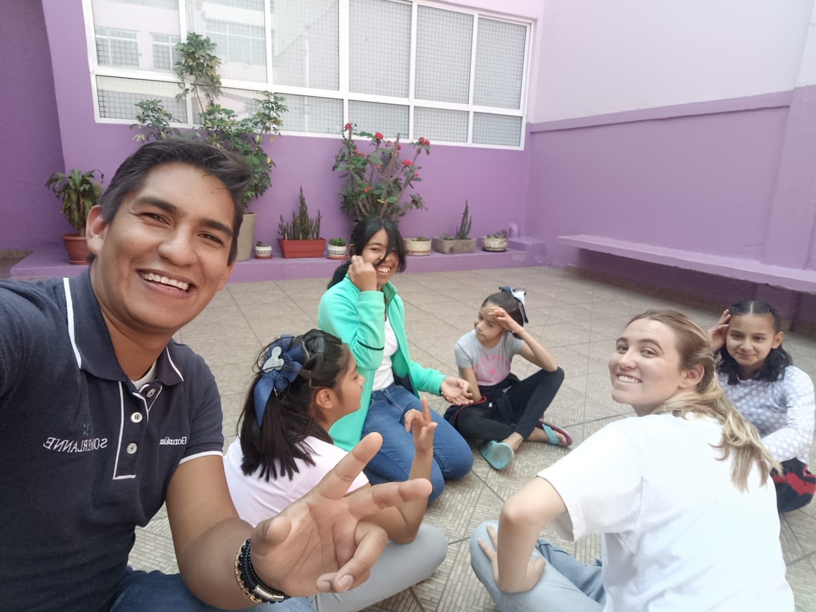 Grupo de personas sonriendo en un patio con paredes violetas, incluyendo adultos y niños sentados en el suelo. Al fondo hay plantas en macetas.