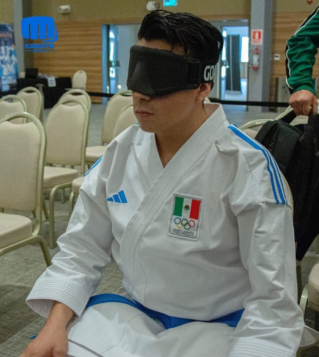 Persona con uniforme de karate y antifaz sentado en una sala con sillas blancas. En el uniforme hay una bandera de México y un logotipo olímpico.