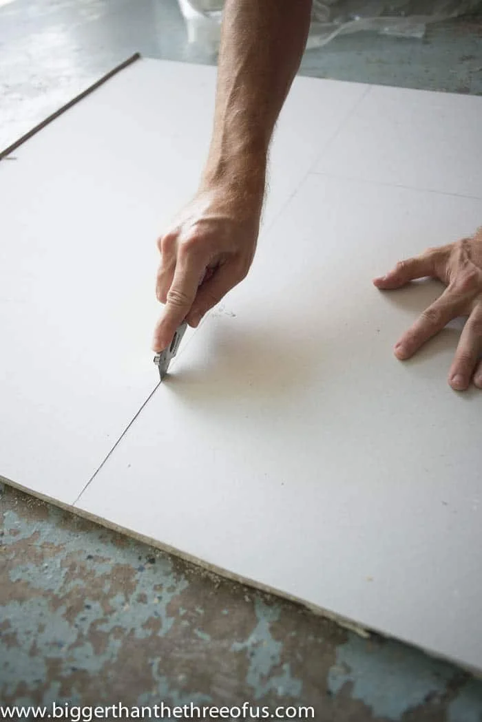 A person is using a utility knife to cut a large white sheet of material, which appears to be foam board or similar, on a work surface.