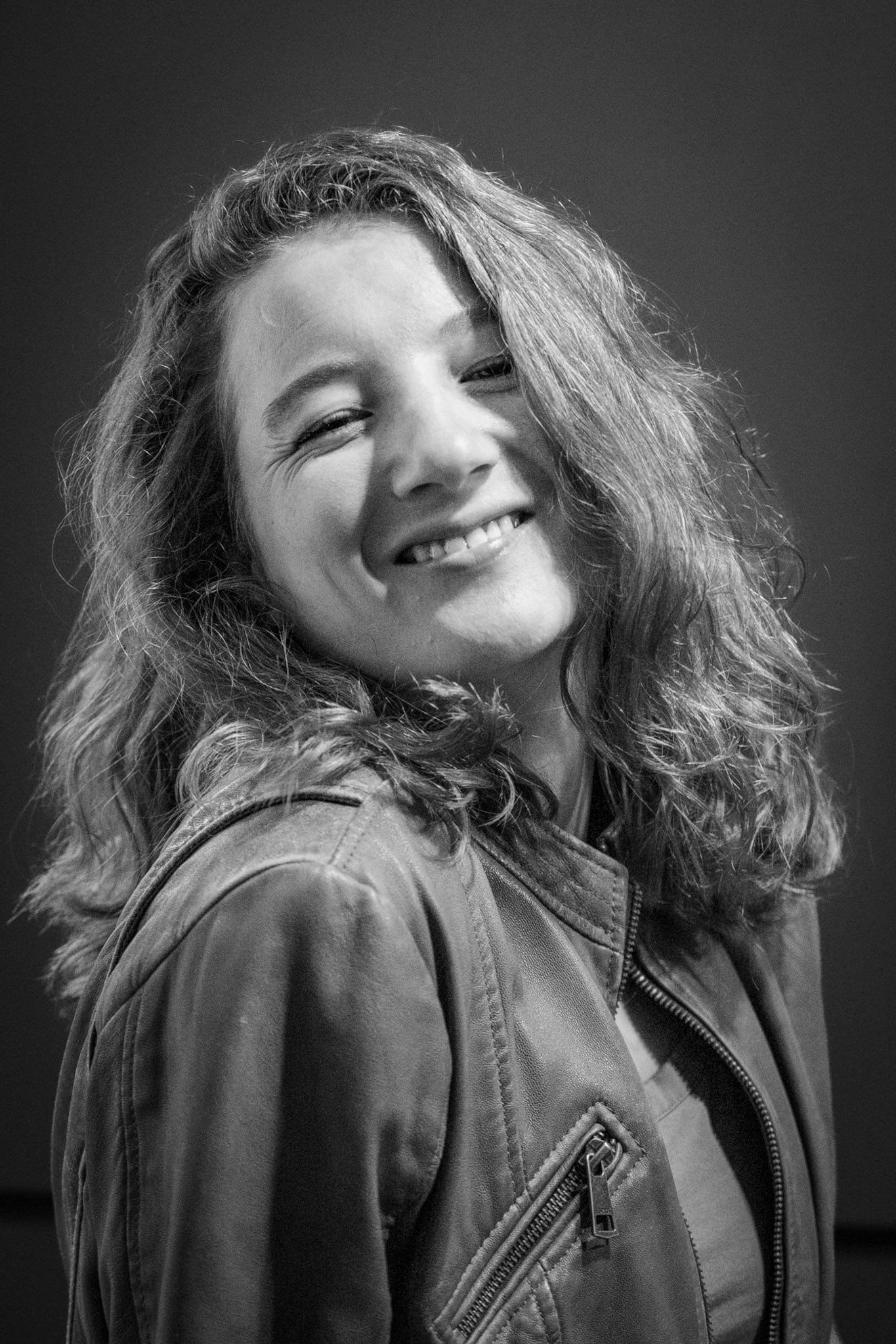 Black and white photo of a smiling woman with curly hair, wearing a jacket with a zipper, against a dark background.