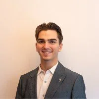 A young man with short brown hair, wearing a gray blazer over a white shirt, smiling and standing against a light-colored wall.