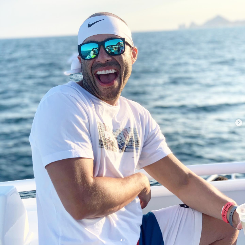 A man smiling and enjoying himself on a boat, wearing sunglasses, a white headband, a white t-shirt, and shorts, with the ocean in the background.