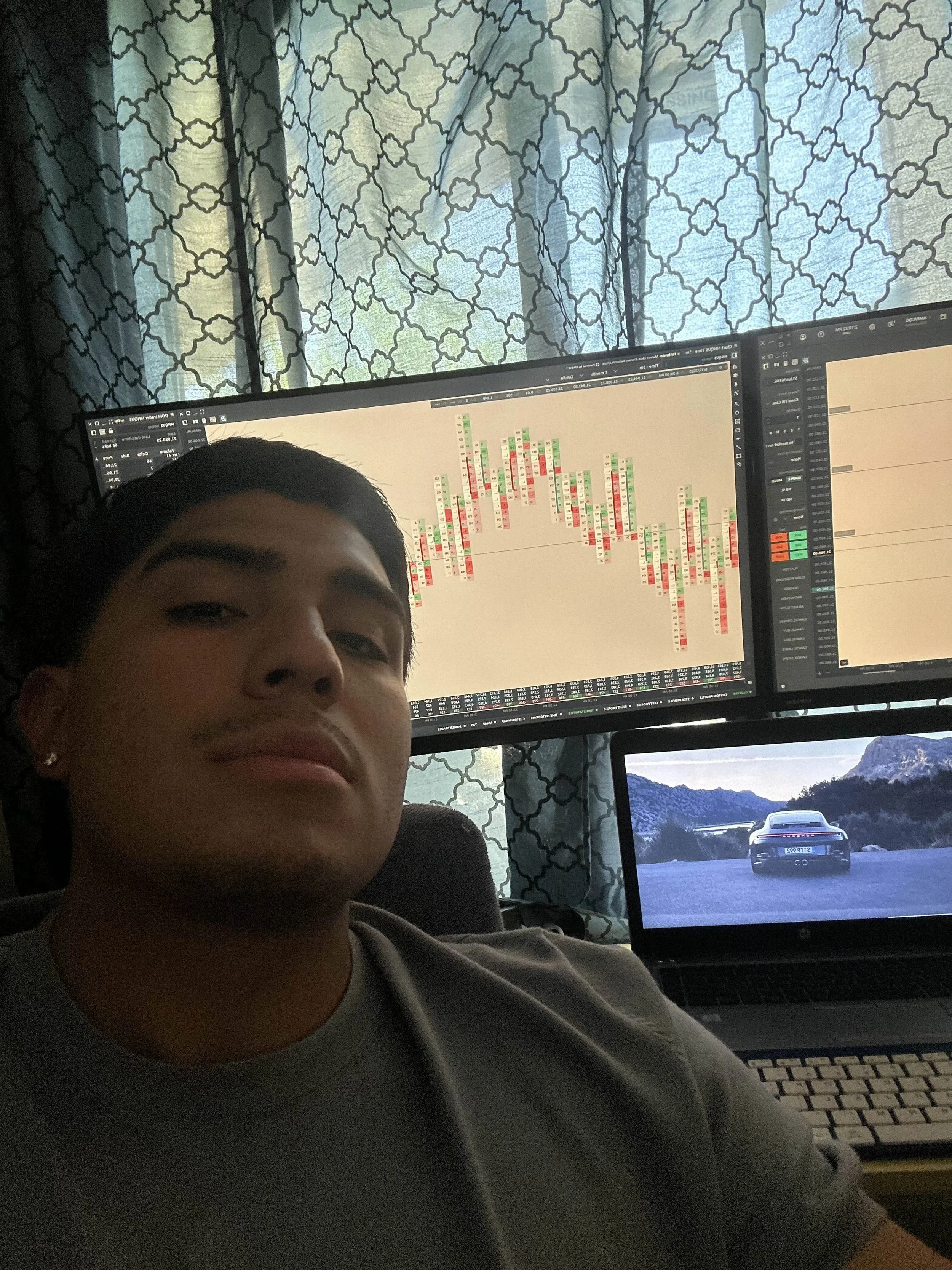 Young man taking a selfie in front of two computer monitors, one showing a colorful bar chart and the other a spreadsheet, with a third screen displaying a sports car on a road in a mountainous area, in a room with patterned curtains.