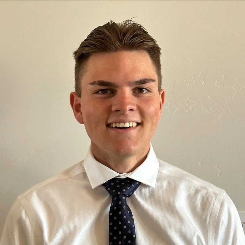 A young man with short, styled brown hair wearing a white dress shirt and a navy blue tie with small white patterns, smiling at the camera, standing against a plain light-colored wall.