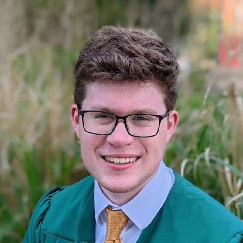 Young man with brown curly hair, glasses, wearing a teal graduation gown and yellow tie, smiling outdoors with greenery in the background.