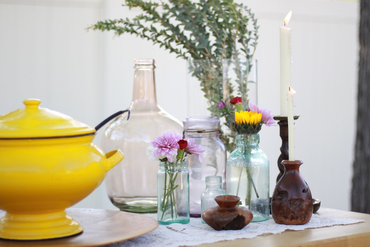 Decorative table arrangement with various glass vases, flowers, a yellow teapot, and candles on a lace cover.