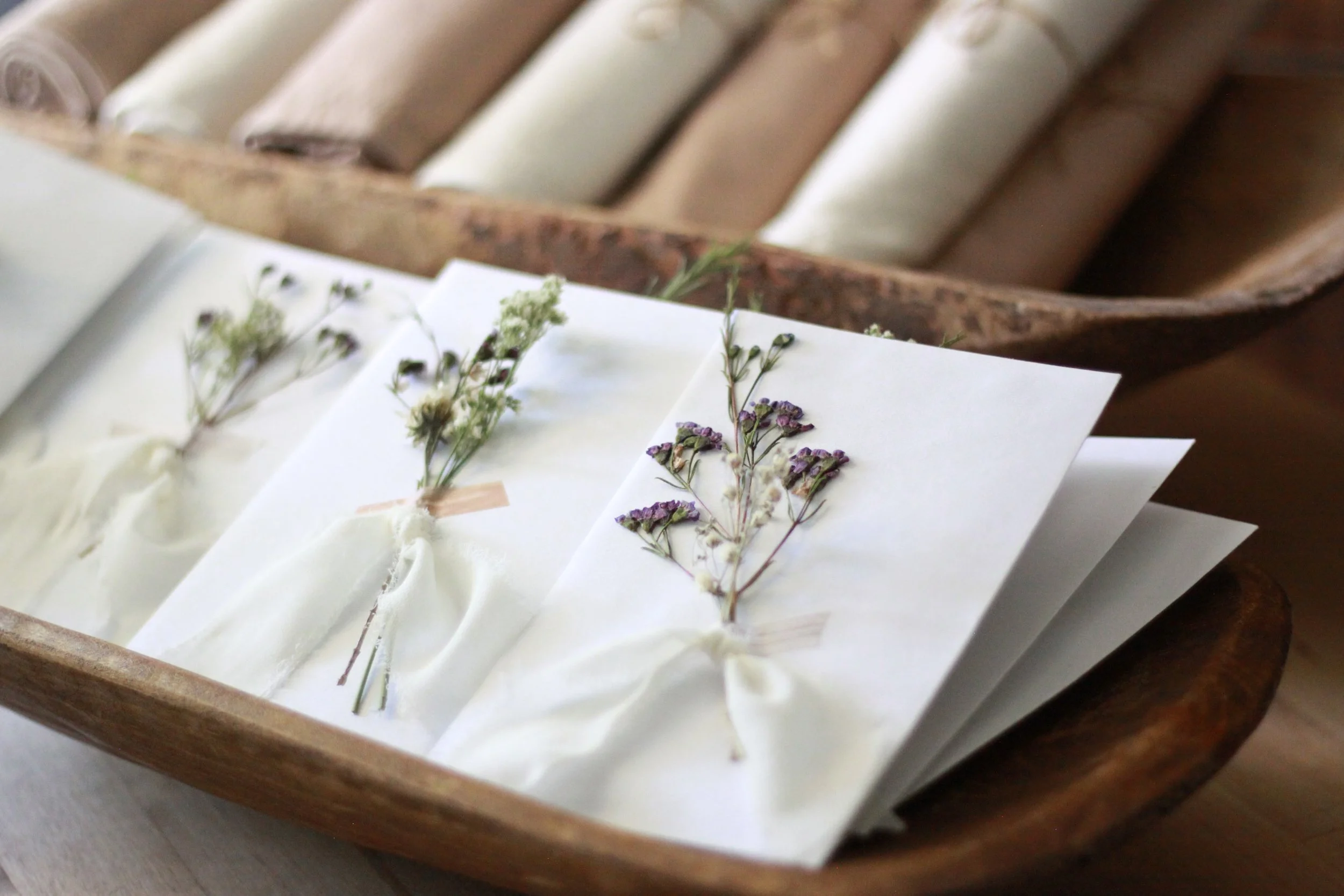 Vintage wooden bowls, hand-tied cloth napkins, and ribbon-wrapped keepsake envelopes create a layered tablescape centered on texture, craftsmanship, and thoughtful experiential event design.
