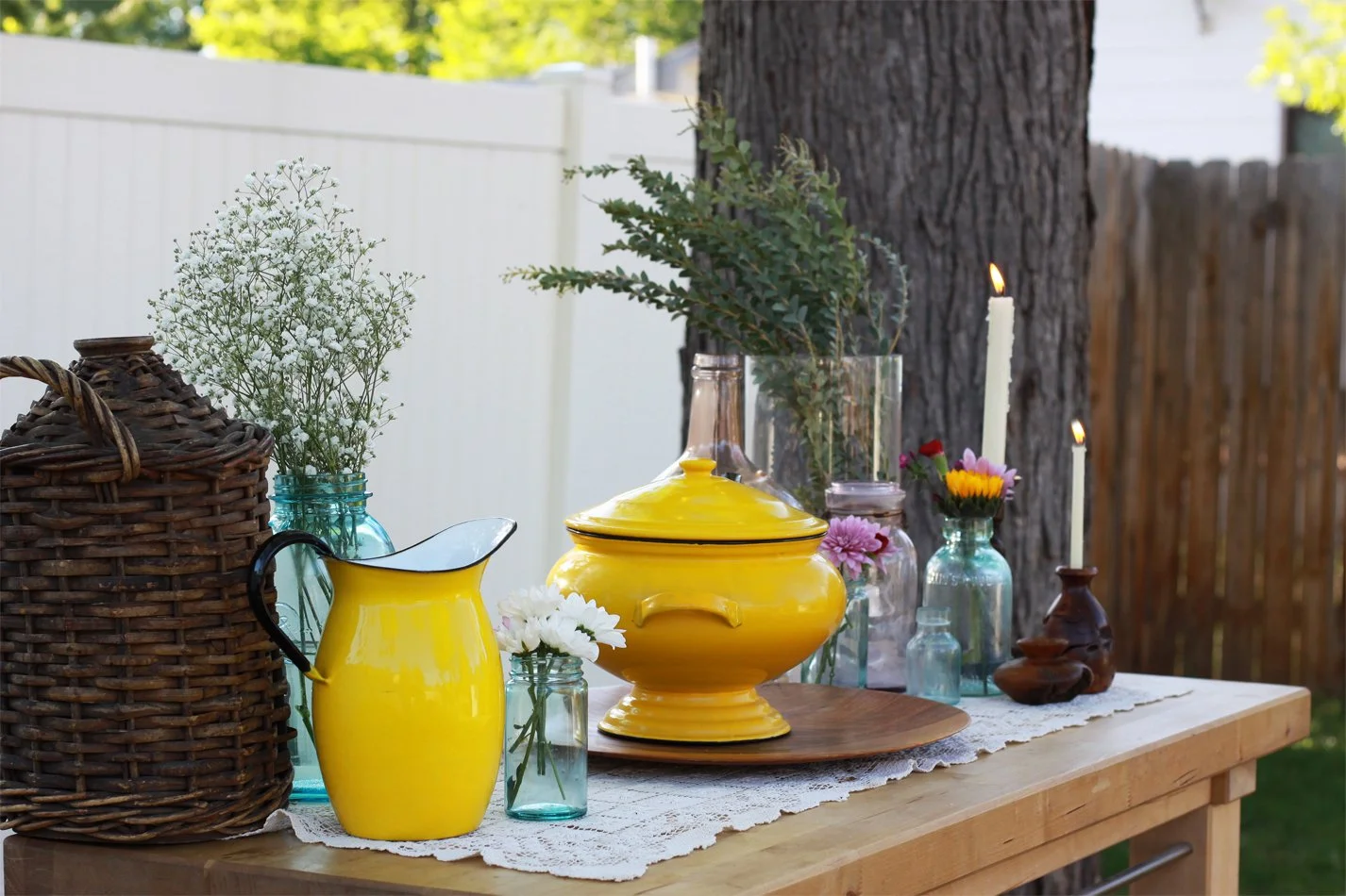 Decorative outdoor table setup with vases of flowers, candles, and a yellow porcelain container under a lace tablecloth.