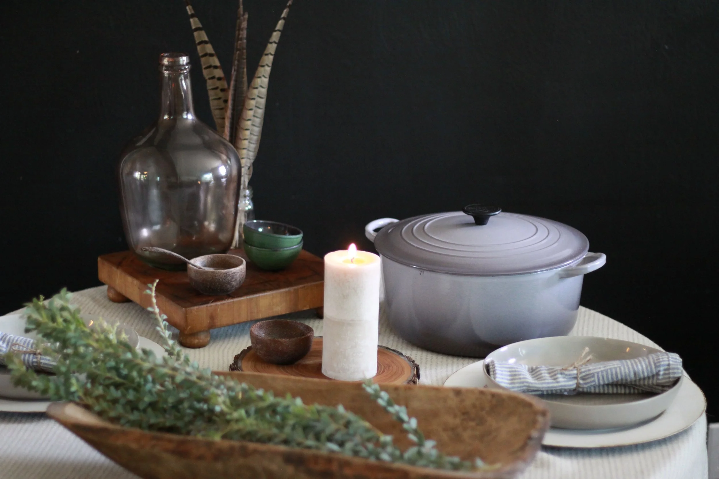 A table set with various kitchen items including a large silver pot, green bowls, small cups, a wooden tray, a white pillar candle, and a white ceramic dish with a lid, against a black background.