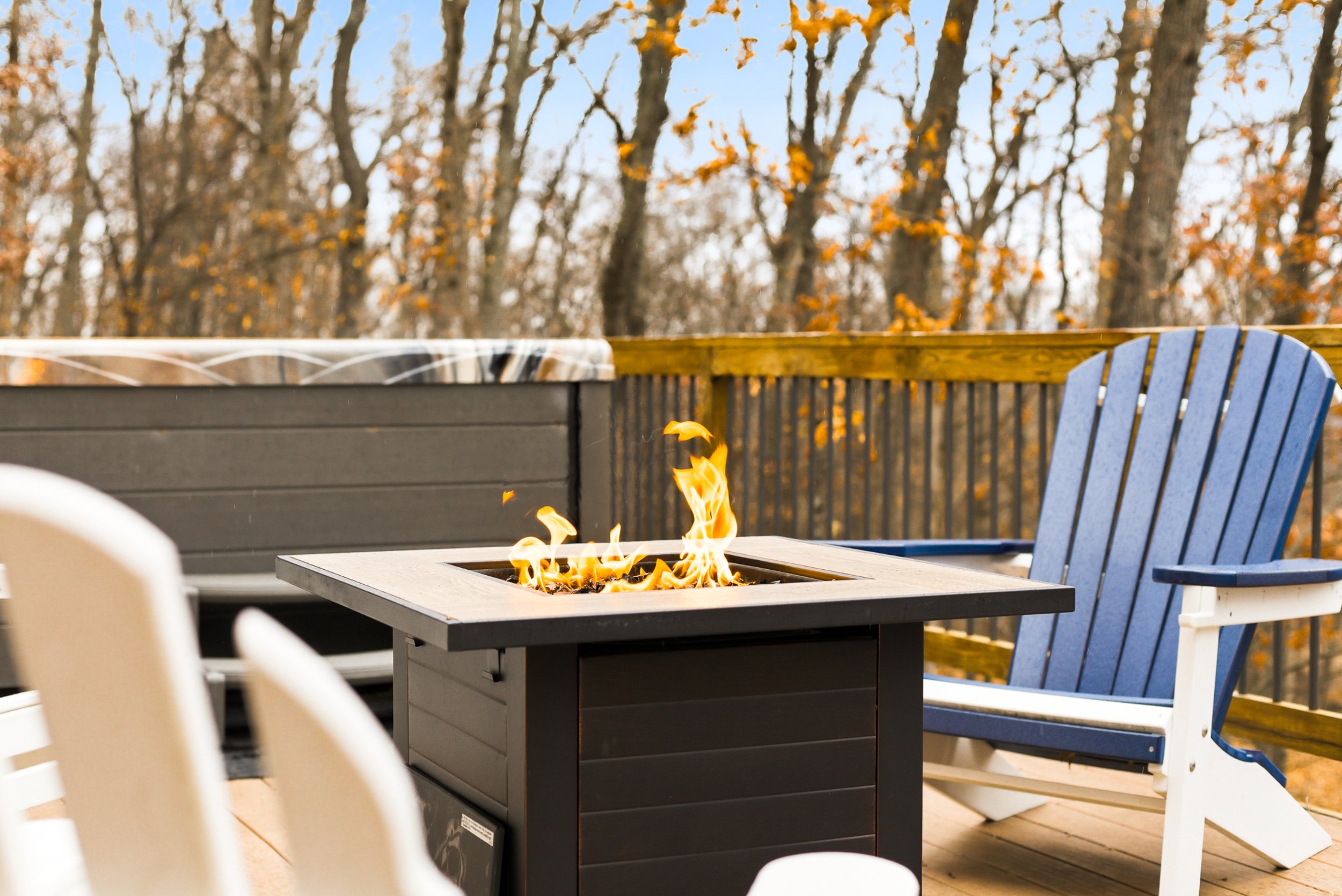Outdoor deck with a fire pit, blue Adirondack chair, and white chairs, surrounded by trees with fall foliage.