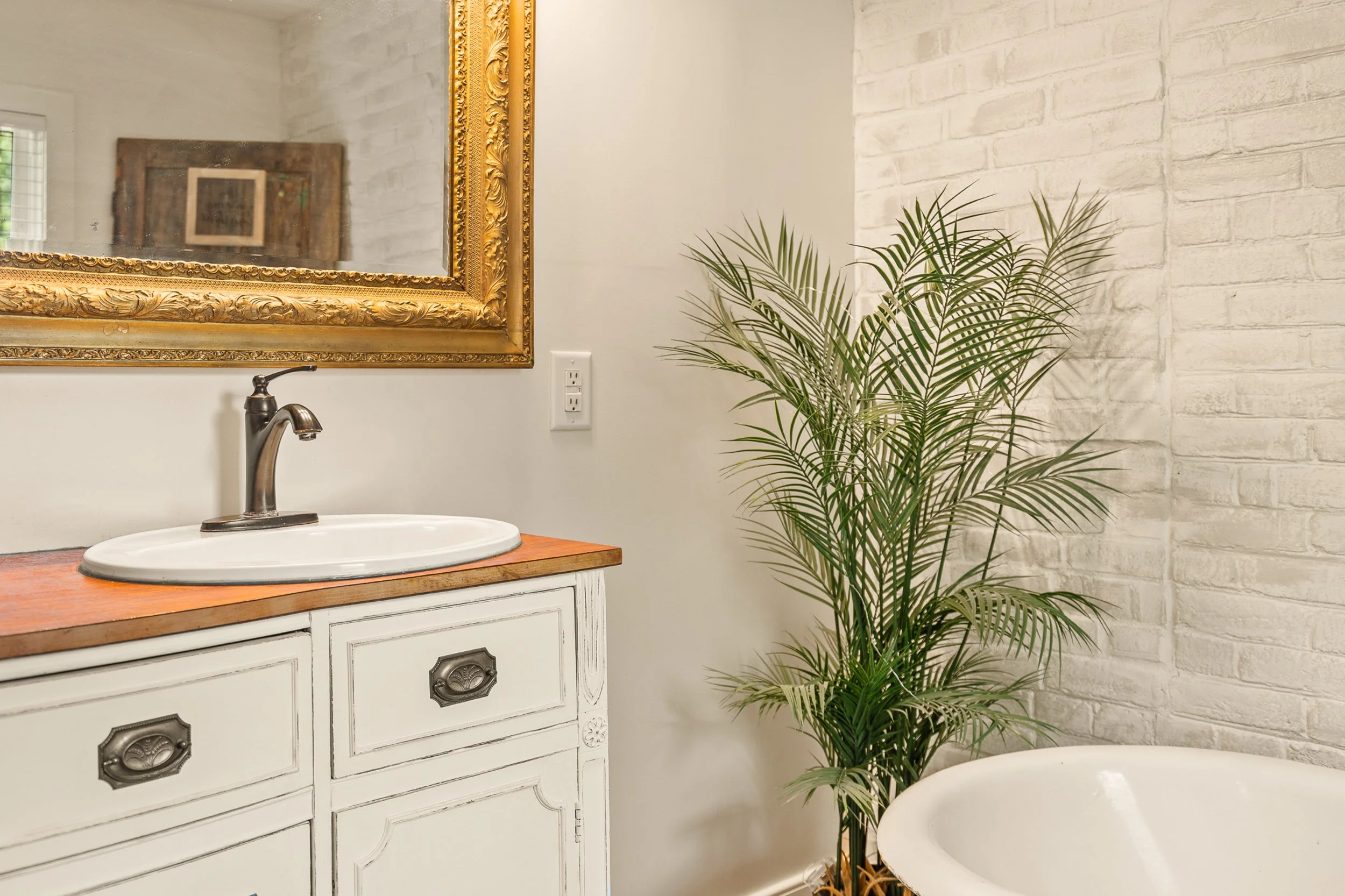 Bathroom with a white vanity, framed mirror, sink and faucet, a potted palm, white brick wall, and a white bathtub.