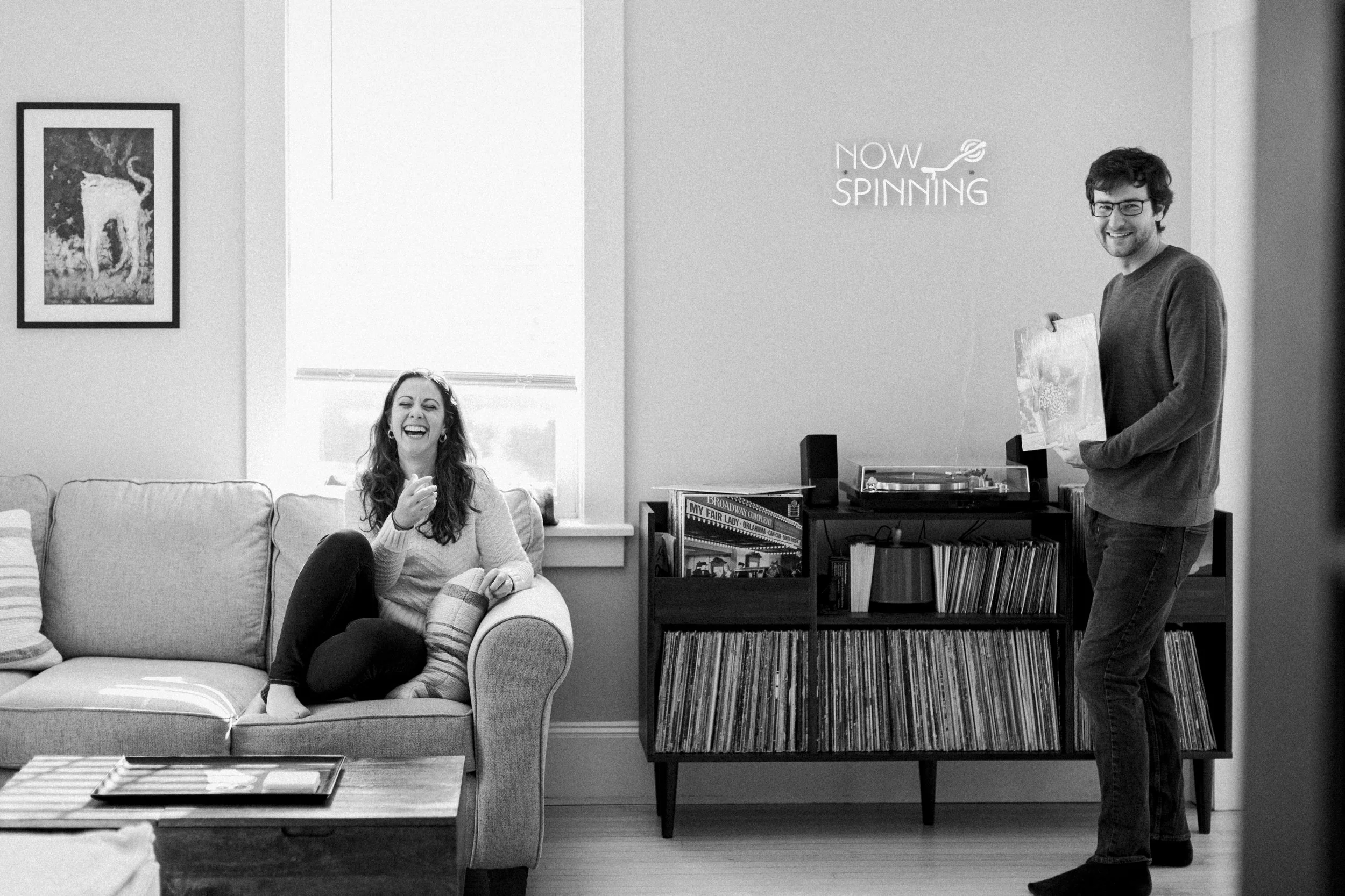 A woman sitting on a sofa laughing, and a man standing on the right holding a record, in a room with a record player and books on a shelf, with a poster on the wall that says 'Now Spinning.'