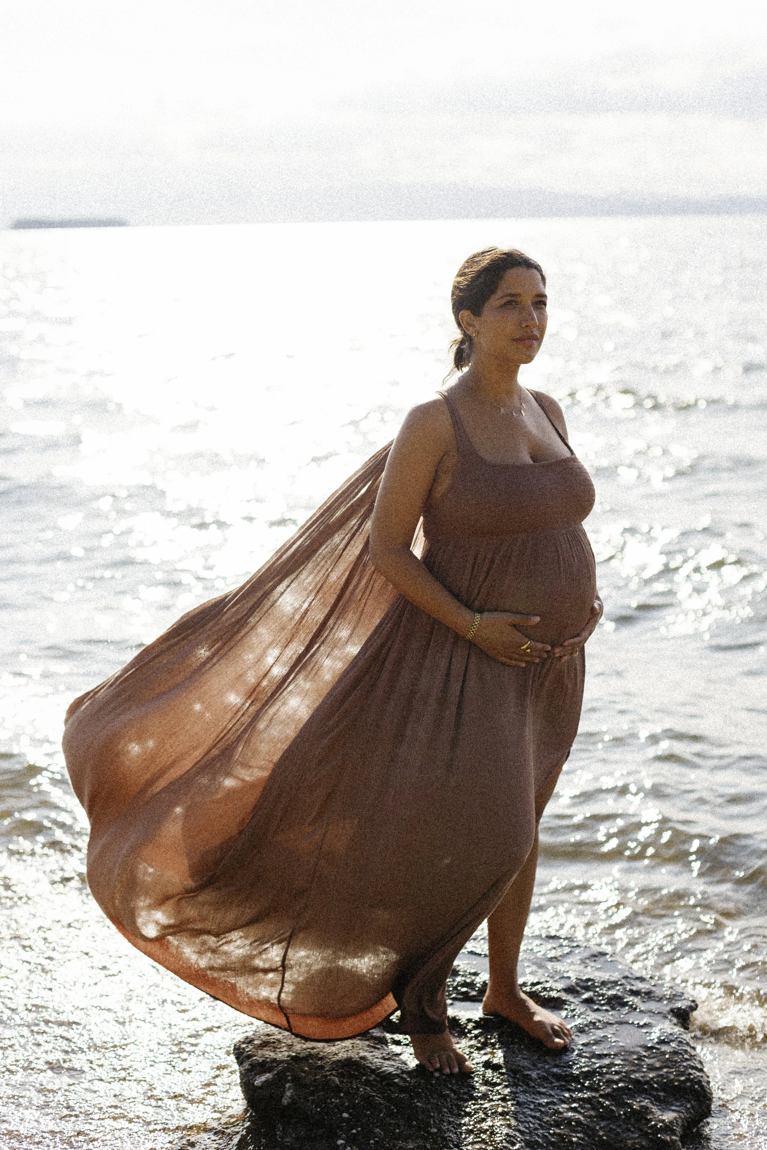 Pregnant woman standing on rocks at the beach, wearing a flowing brown dress and holding her belly, with the ocean and sunlight in the background.