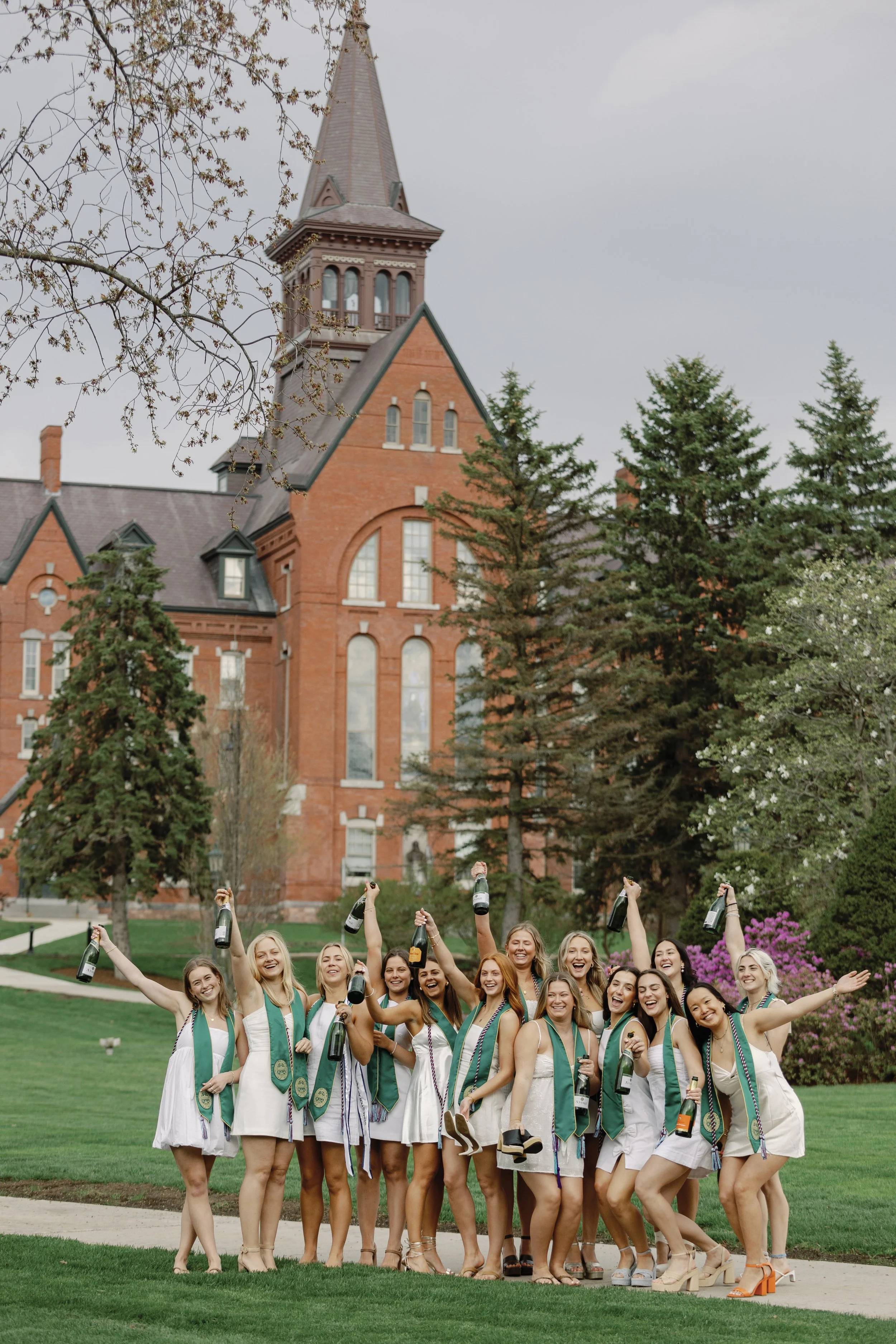 A group of women in white dresses celebrating outdoors with champagne bottles, standing on a green lawn in front of a brick building with a tall steeple.