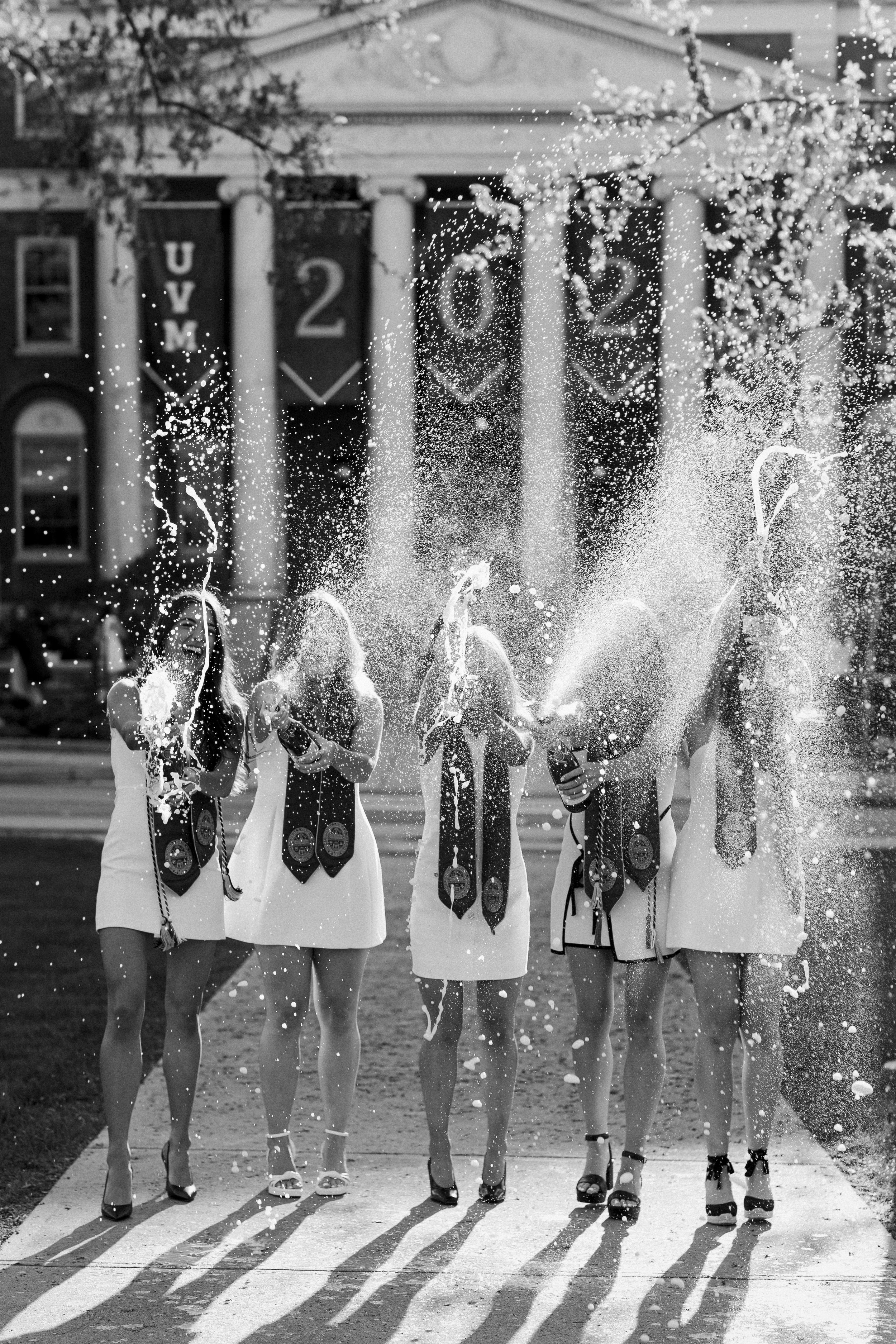 Five women wearing white dresses and medals celebrating New Year's Eve 2022, spraying champagne in front of a building with banners that read VM 2022