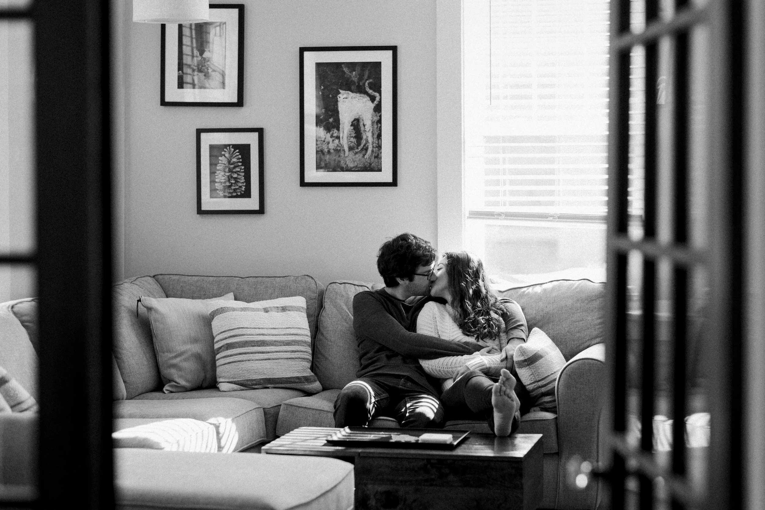 A couple sharing a kiss on a couch in a living room, seen through a doorway, with framed artwork on the wall and sunlight coming through blinds.
