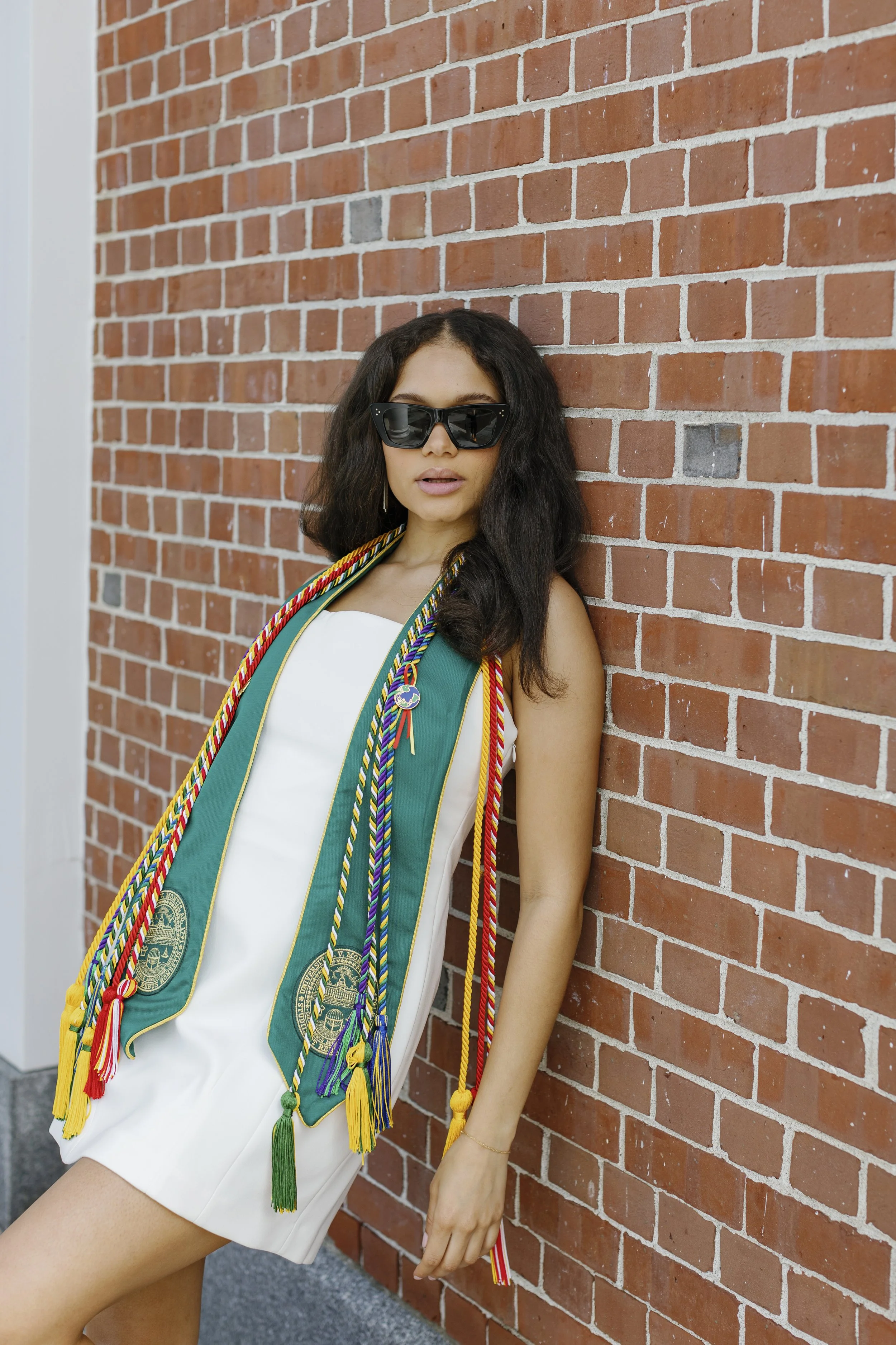 A woman wearing sunglasses and a white dress stands against a brick wall, with a graduation stole around her neck.