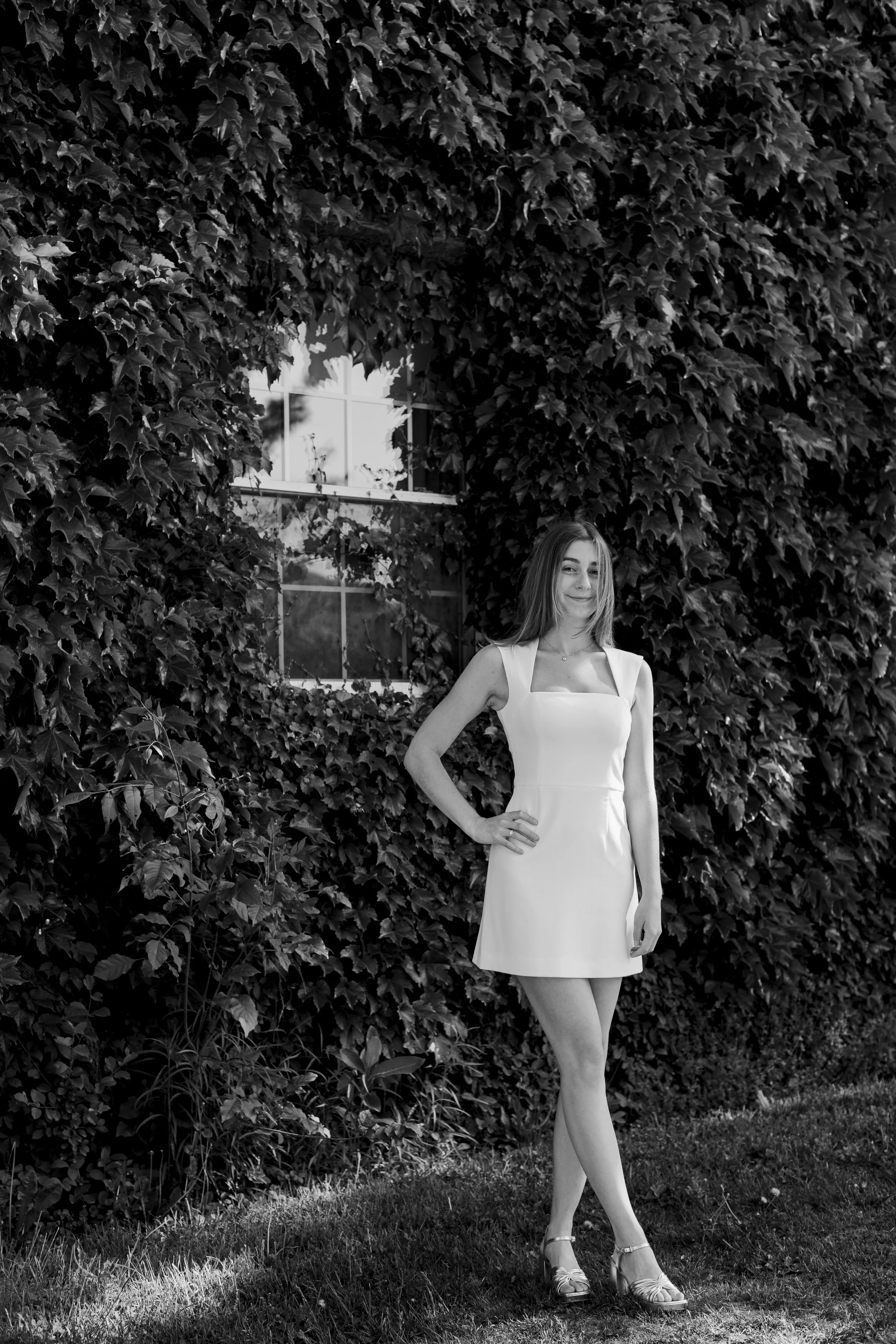 A woman in a light-colored dress and sandals standing outside in front of a wall covered with dense ivy.