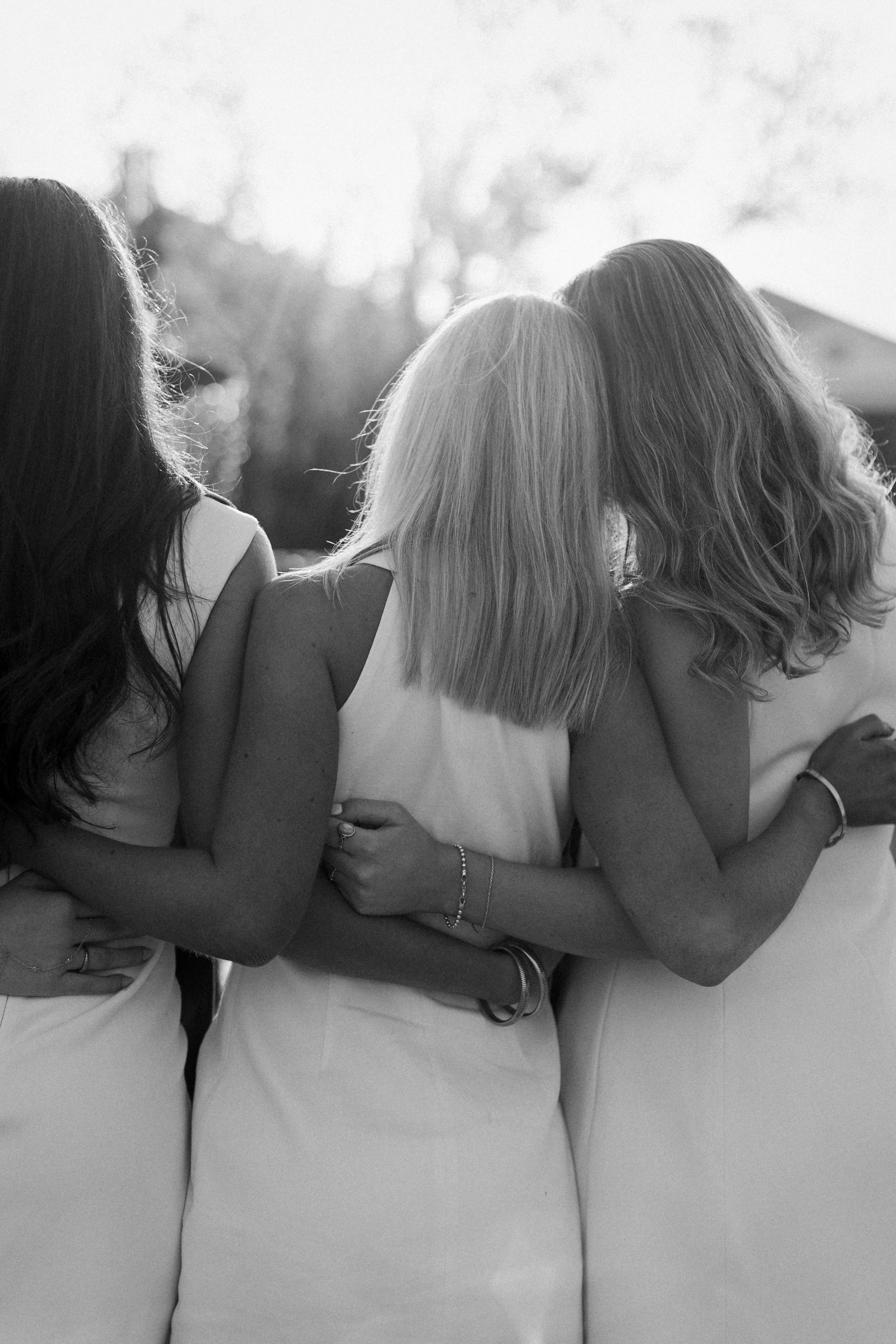 Three women standing outdoors with their arms around each other, photographed from the back, black and white