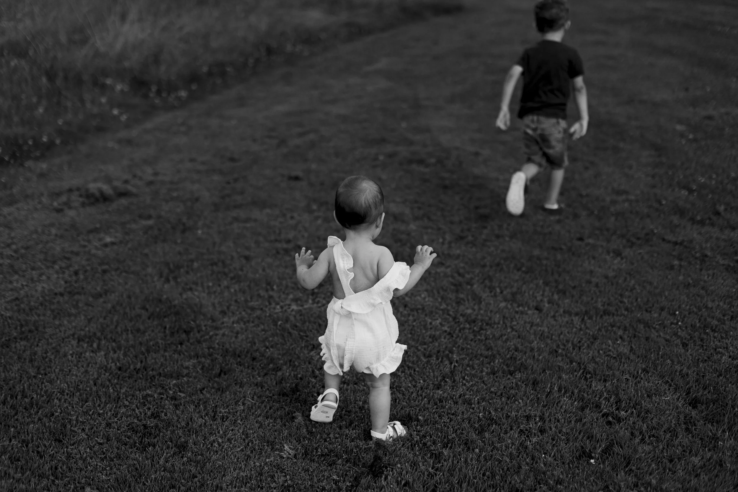 A young girl and a boy running on a grassy field, seen from behind, with the girl in a light dress and the boy in a black t-shirt and shorts.