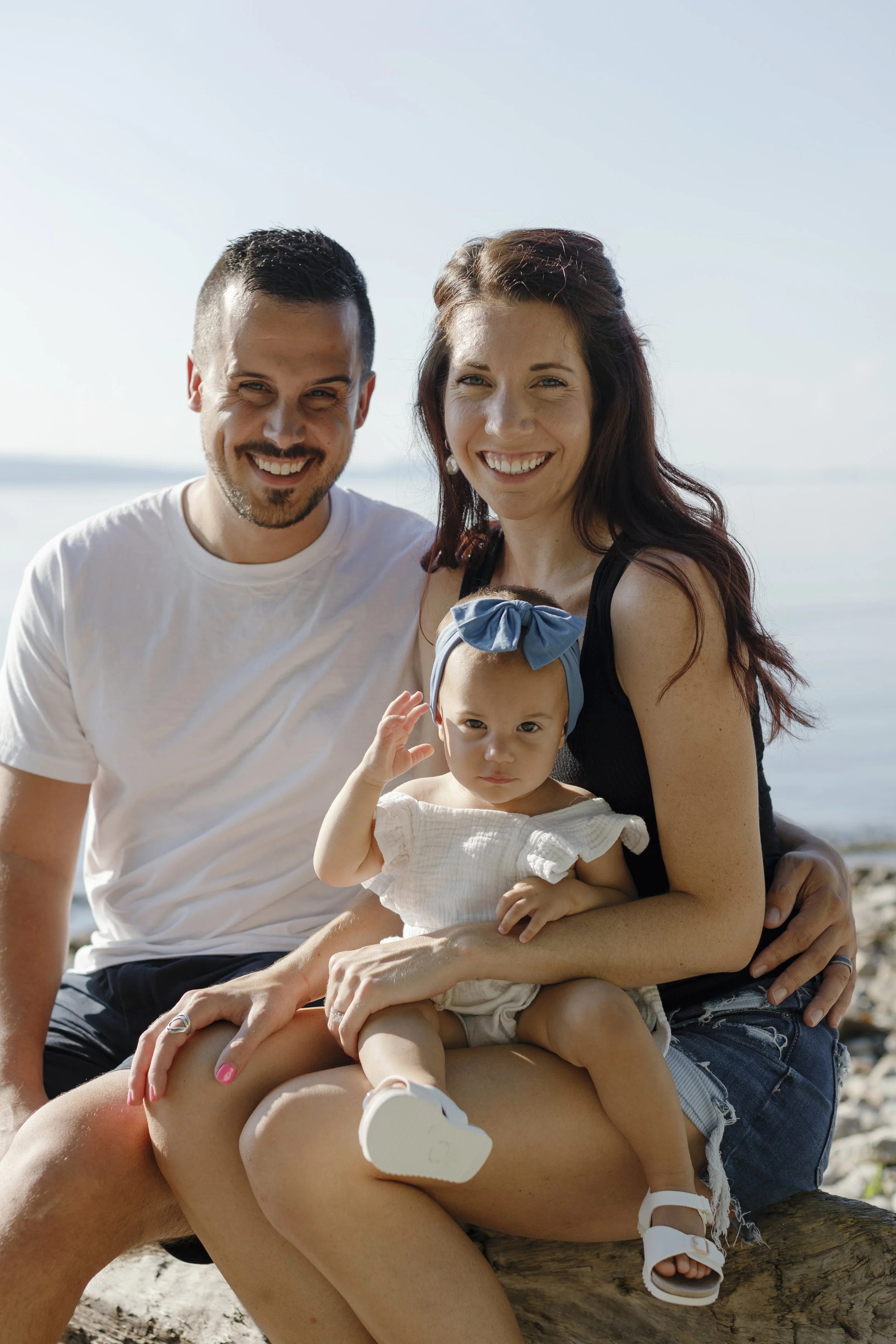 A family of three, a man, a woman, and a young girl, smiling and sitting on a log at the beach.