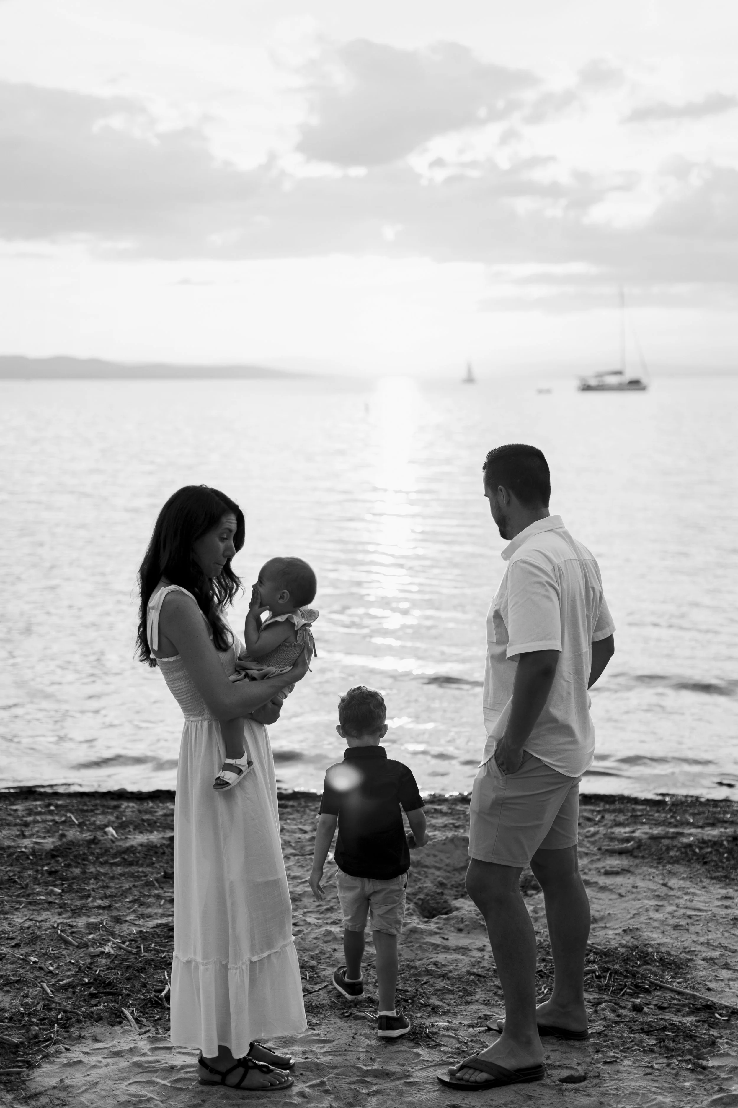 A family of four stands on the beach at sunset, with a mother holding a baby, a father standing nearby, and two young boys walking towards the water, with sailboats in the background.