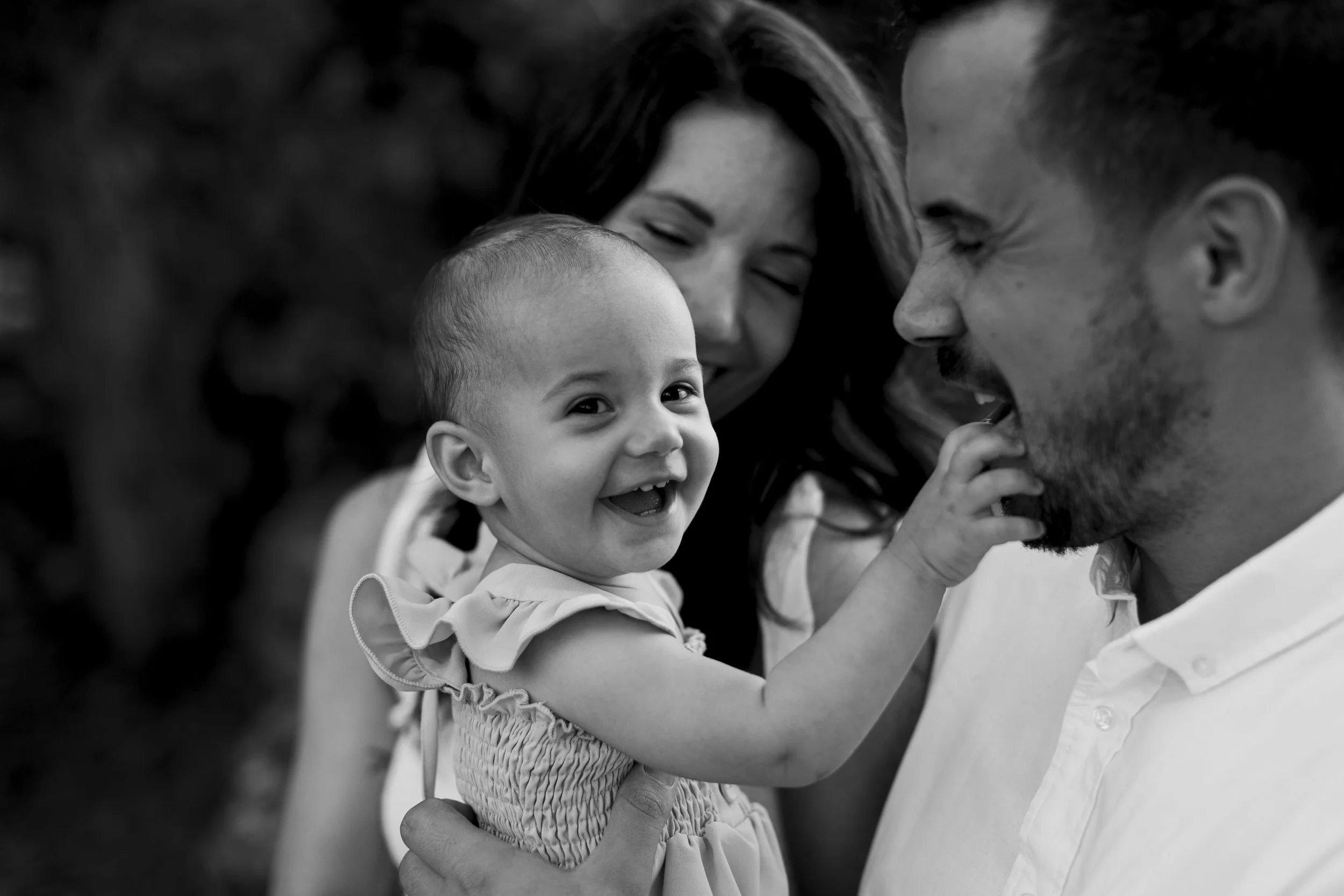 A happy young girl touching her father's face while his partner looks on with a smile in the background, all outdoors.
