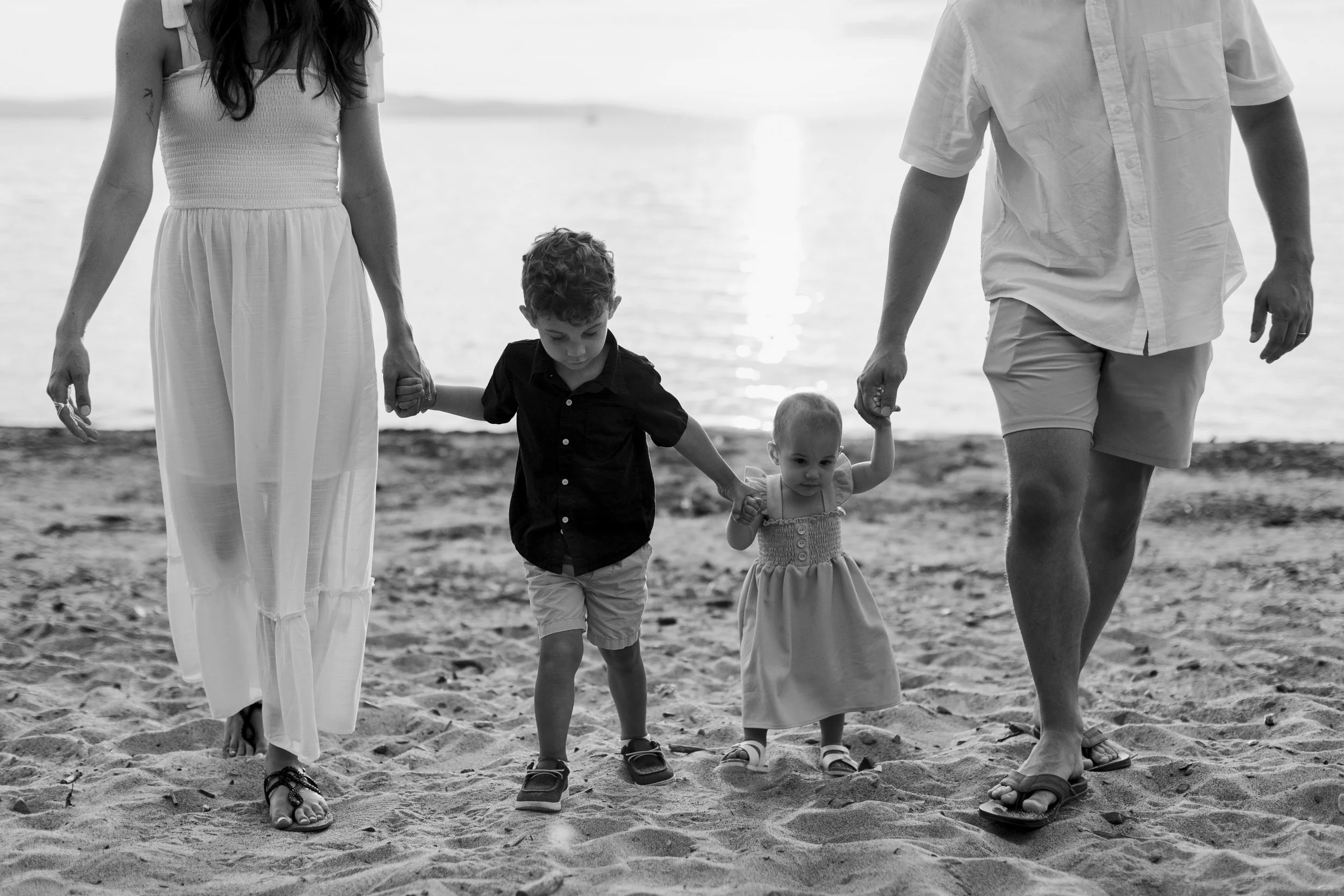 A family of four walking on a sandy beach, holding hands, with water in the background. The parents are on both sides, an older child in the middle holding hands with both parents, and a younger child next to the mother. The image is in black and whi