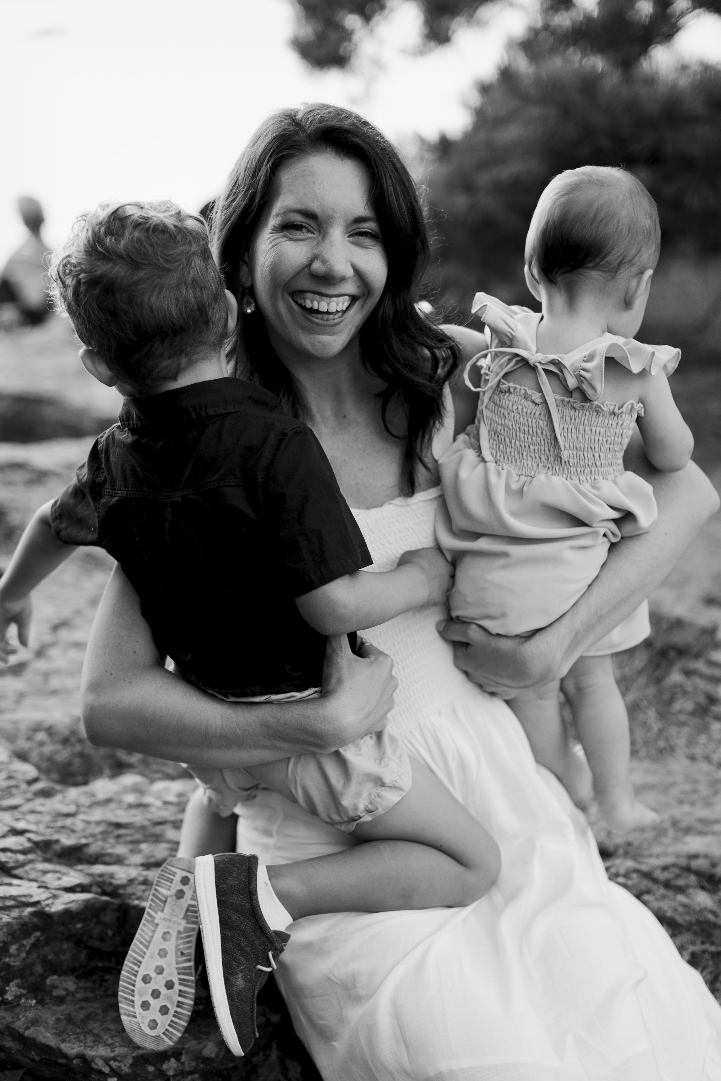A woman smiling while holding two young children outdoors, black and white photo.