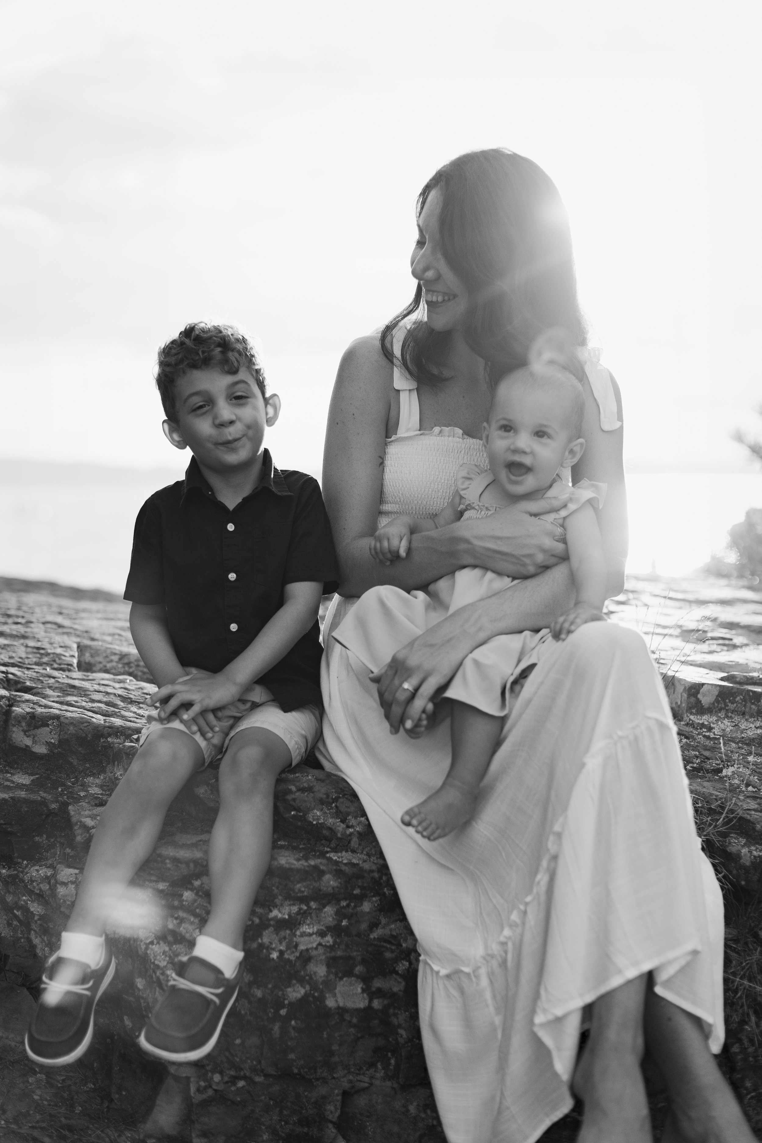 A woman sitting on a stone ledge with two children, one boy and one baby girl, outdoors during the daytime. The woman is smiling and holding the baby girl, while the boy sits beside her. The photo is in black and white.