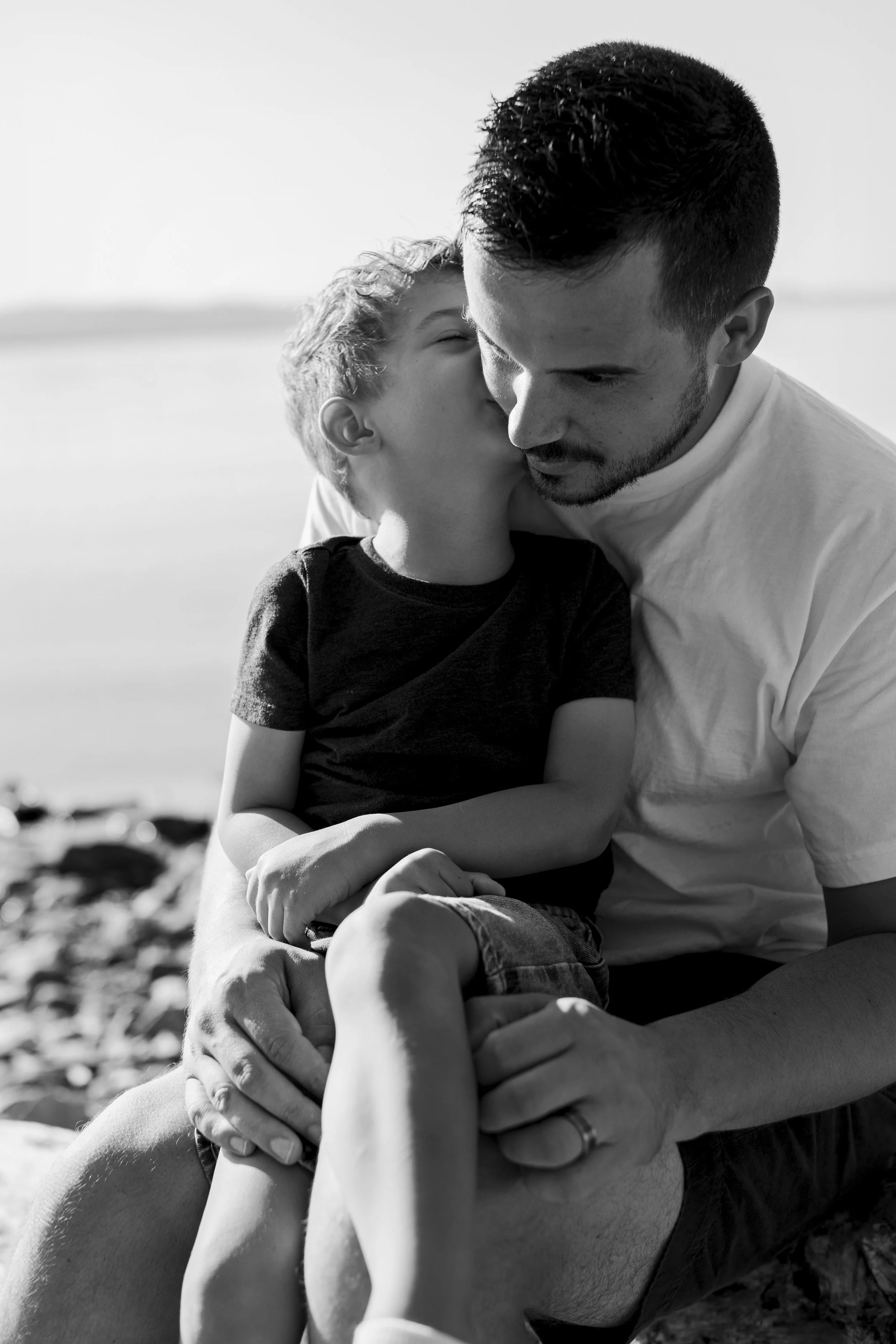 A young boy kissing a man on the cheek while sitting outdoors near water.