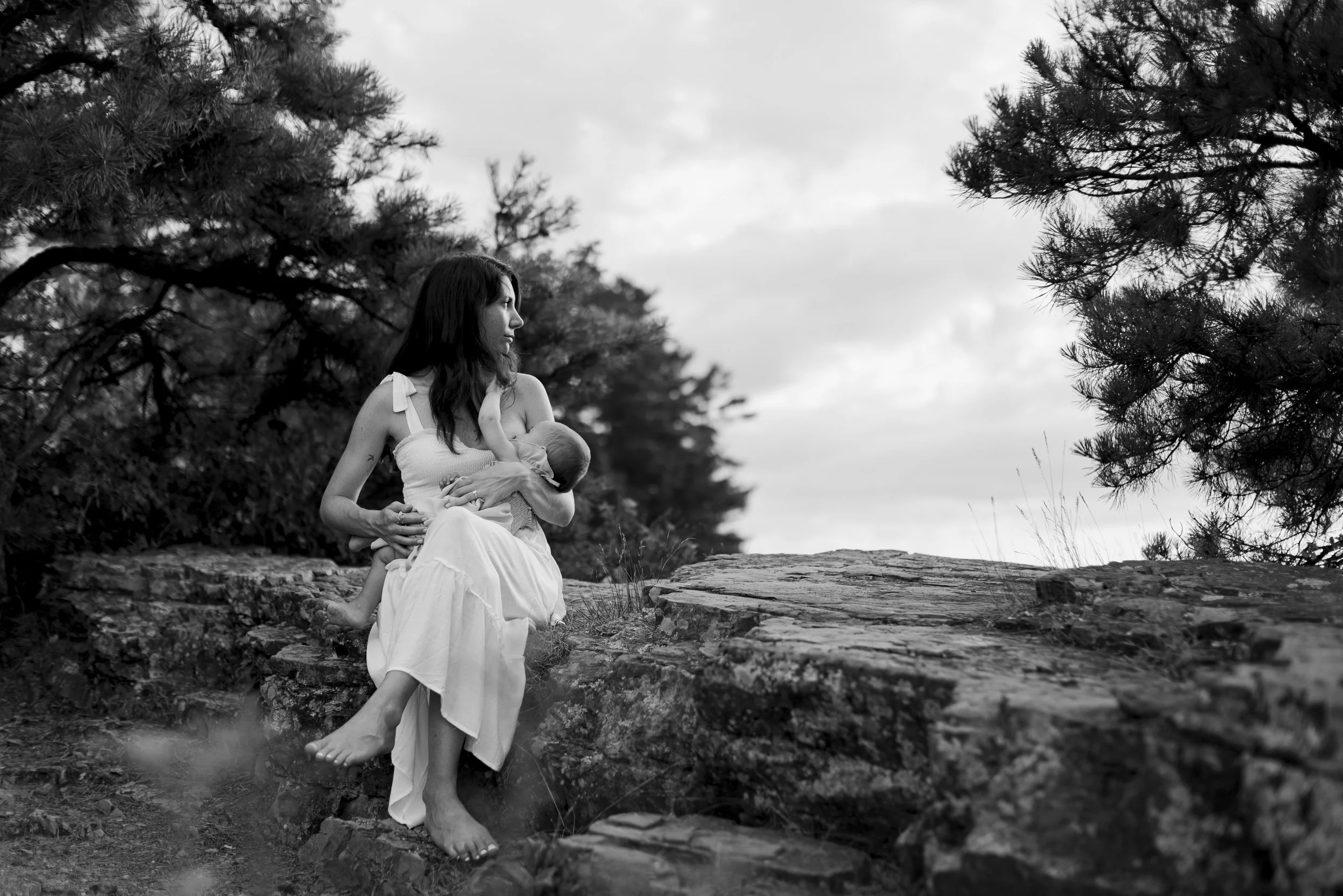 Black and white photograph of a woman sitting on a rock outdoors, breastfeeding a baby. The woman is wearing a long, sleeveless dress with shoulder bows. There are trees and cloudy sky in the background.