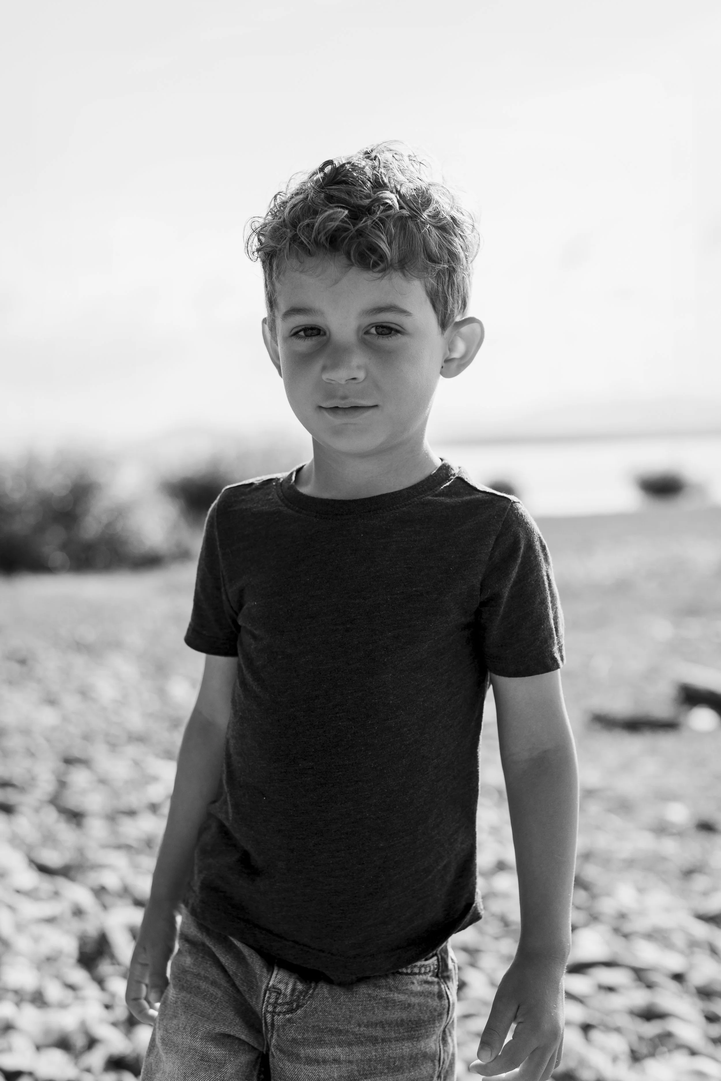 A black and white photo of a young boy with curly hair standing outdoors on a blurred background.