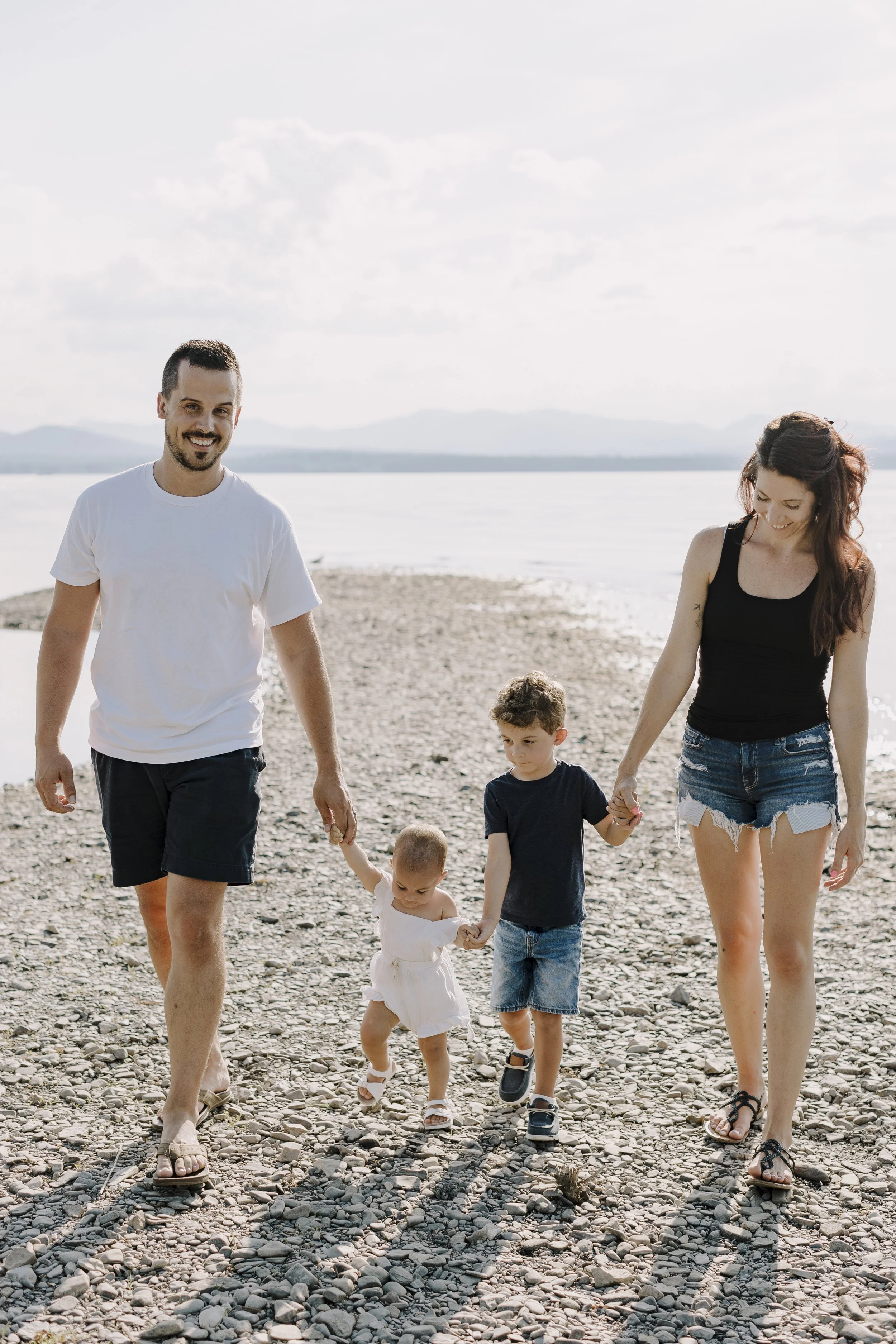 A family of four, including two adults and two young children, walking hand-in-hand on a rocky beach near a body of water with mountains in the background. The man and woman are smiling and wearing casual summer clothes.