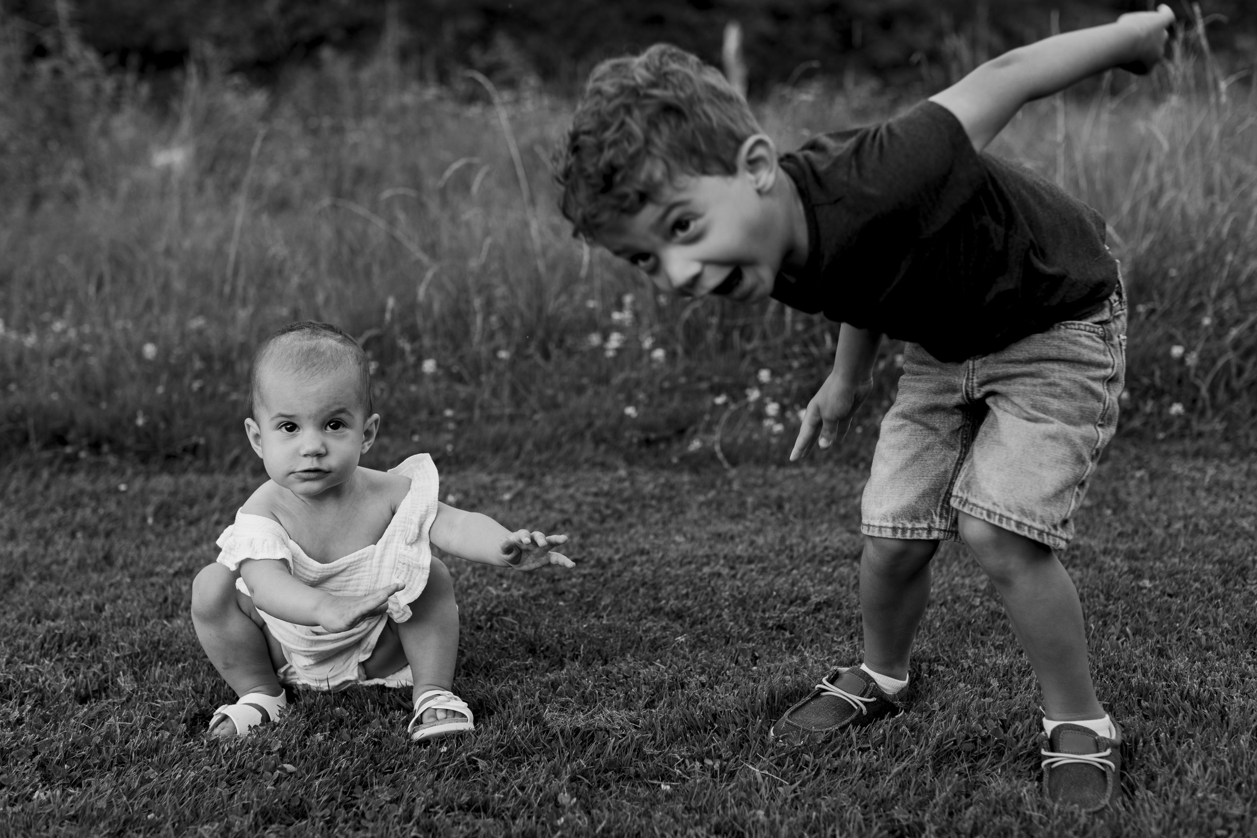 Two young children playing outdoors on grass: a baby girl squatting and looking at the camera, and a boy bending down with his tongue out, smiling.