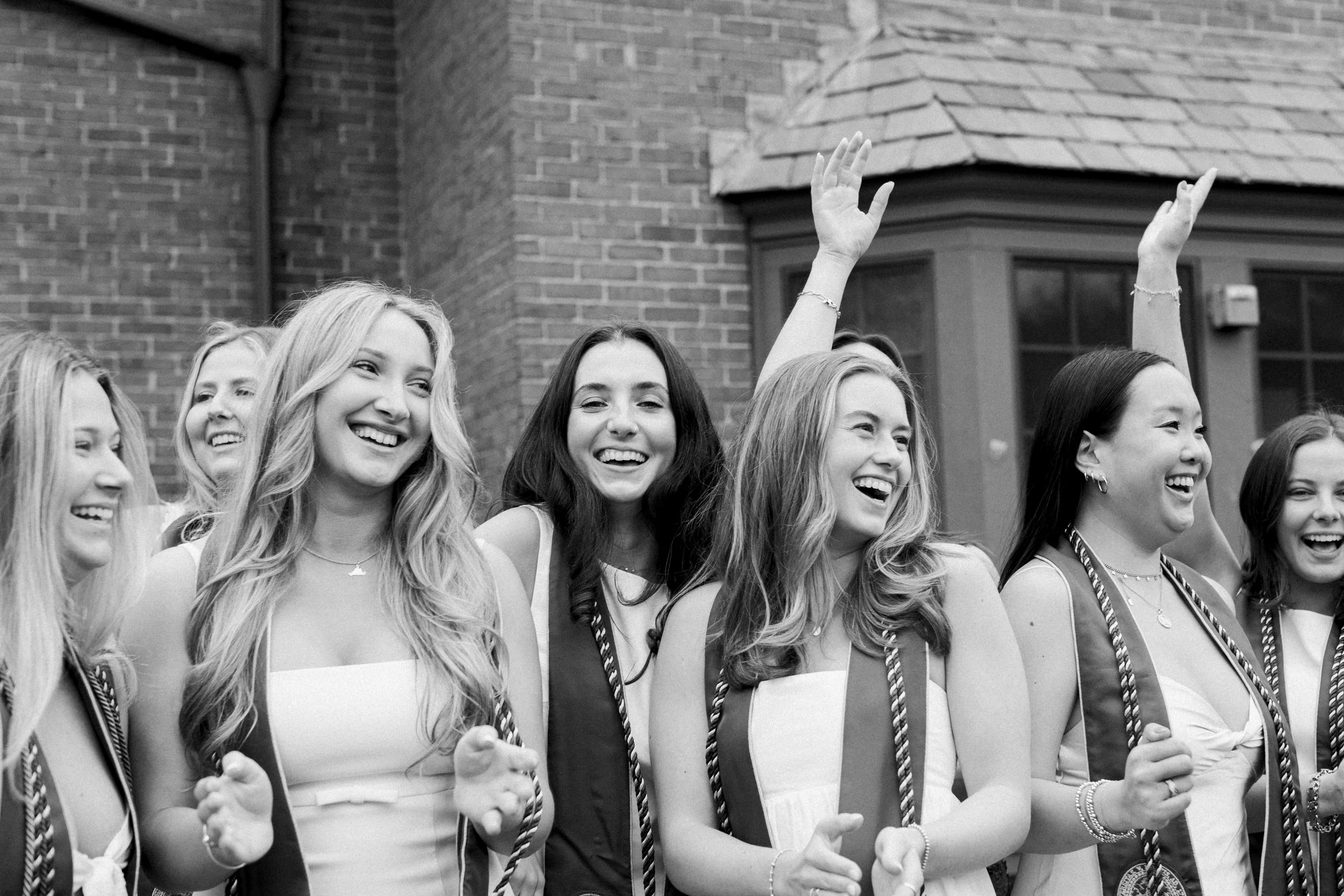 Black and white photo of a group of young women wearing graduation gowns and caps, smiling and celebrating outdoors.