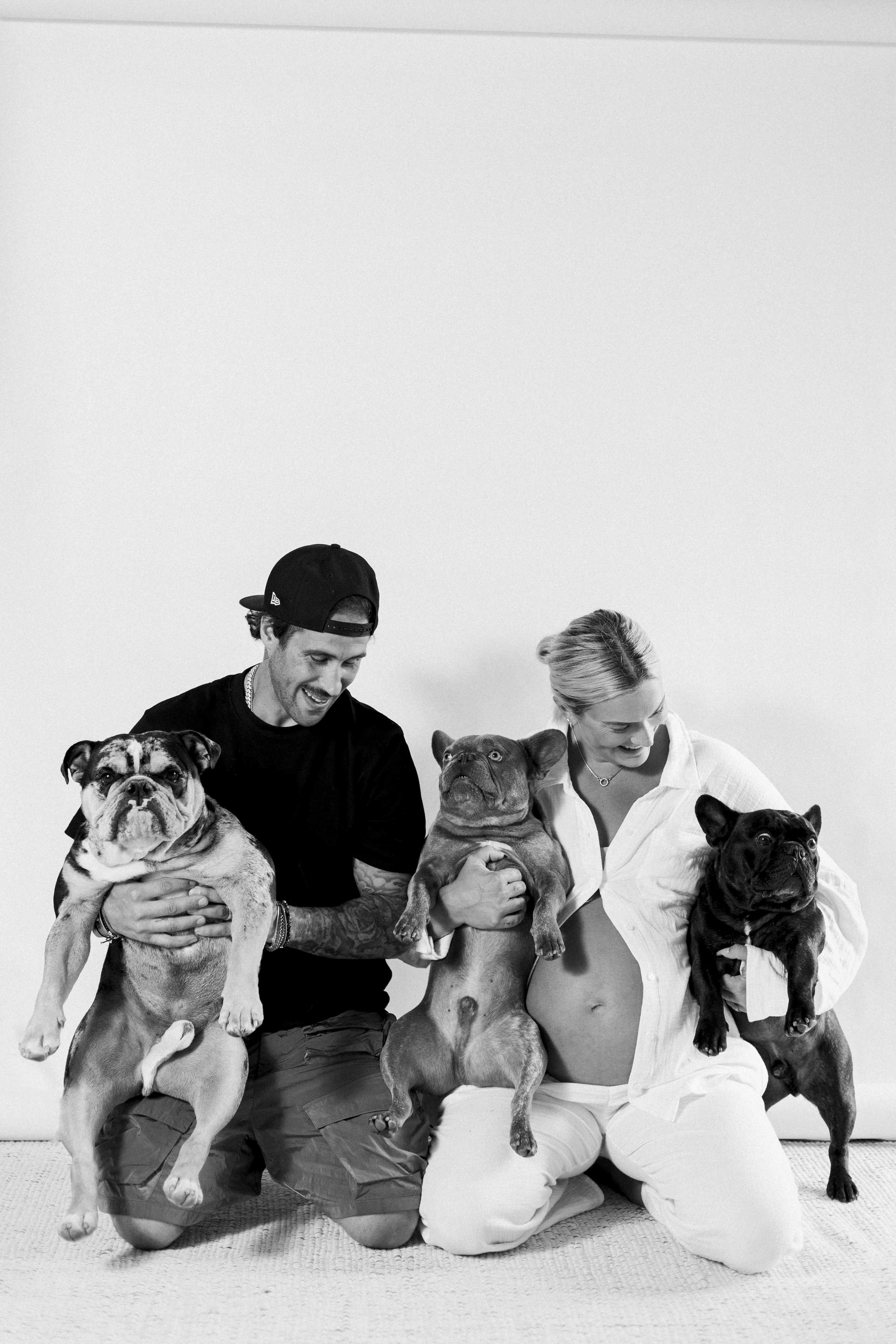A black-and-white photo of a smiling man and woman holding three French bulldogs, against a plain white background.