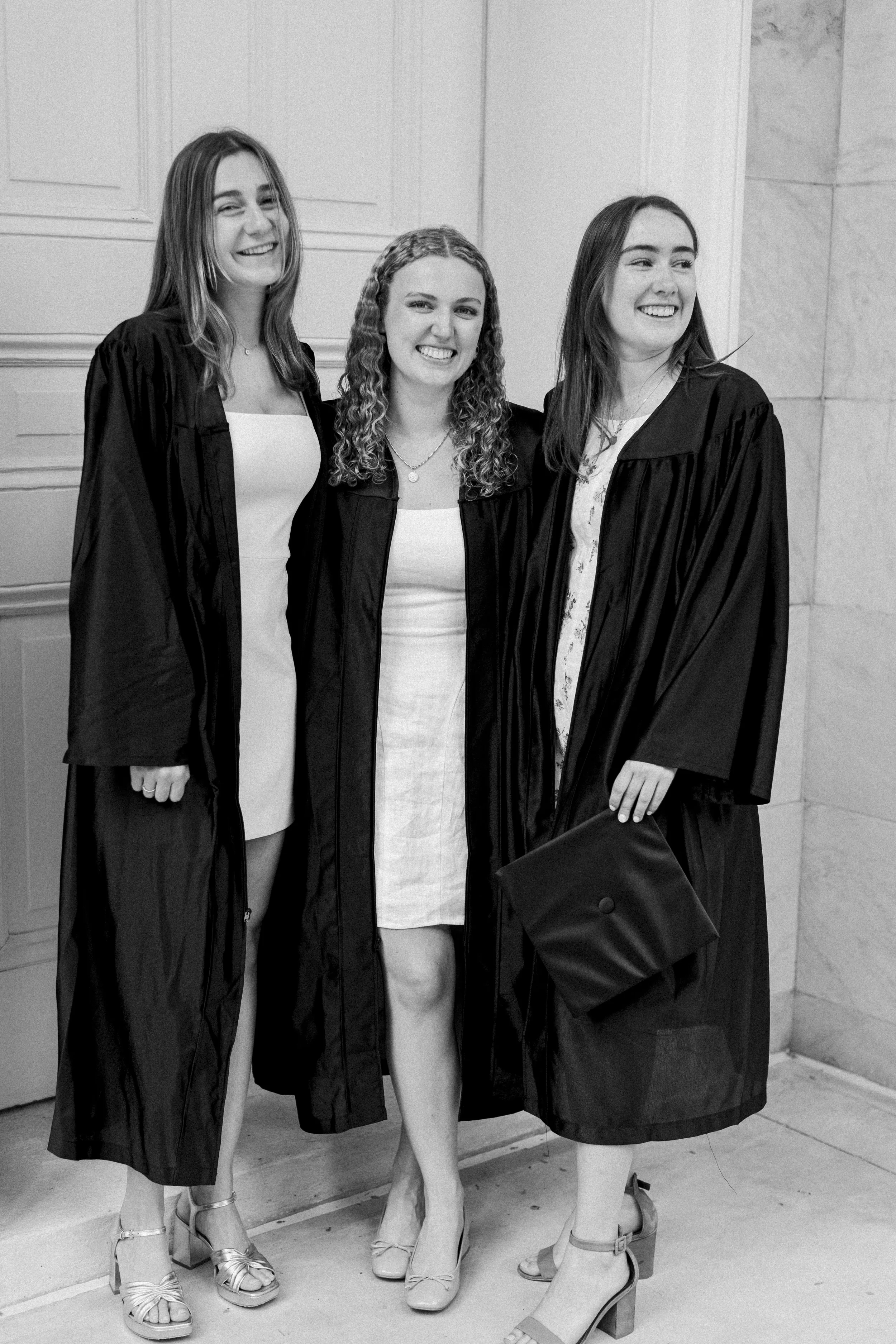 Three young women in graduation gowns and heels standing together, smiling, at a graduation ceremony in black and white.