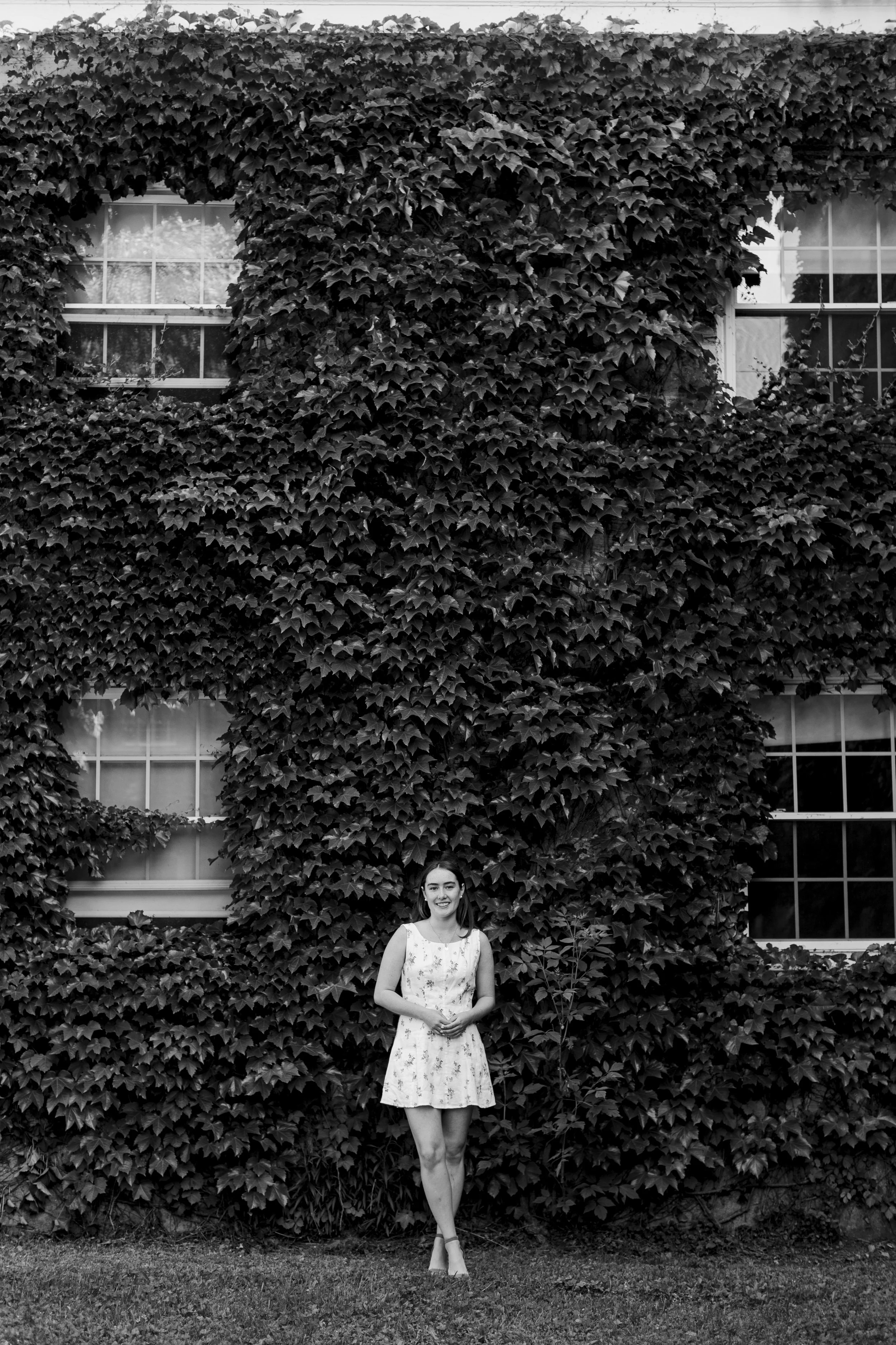 A young woman in a sleeveless, knee-length floral dress standing on grass in front of a large wall covered in ivy with windows.