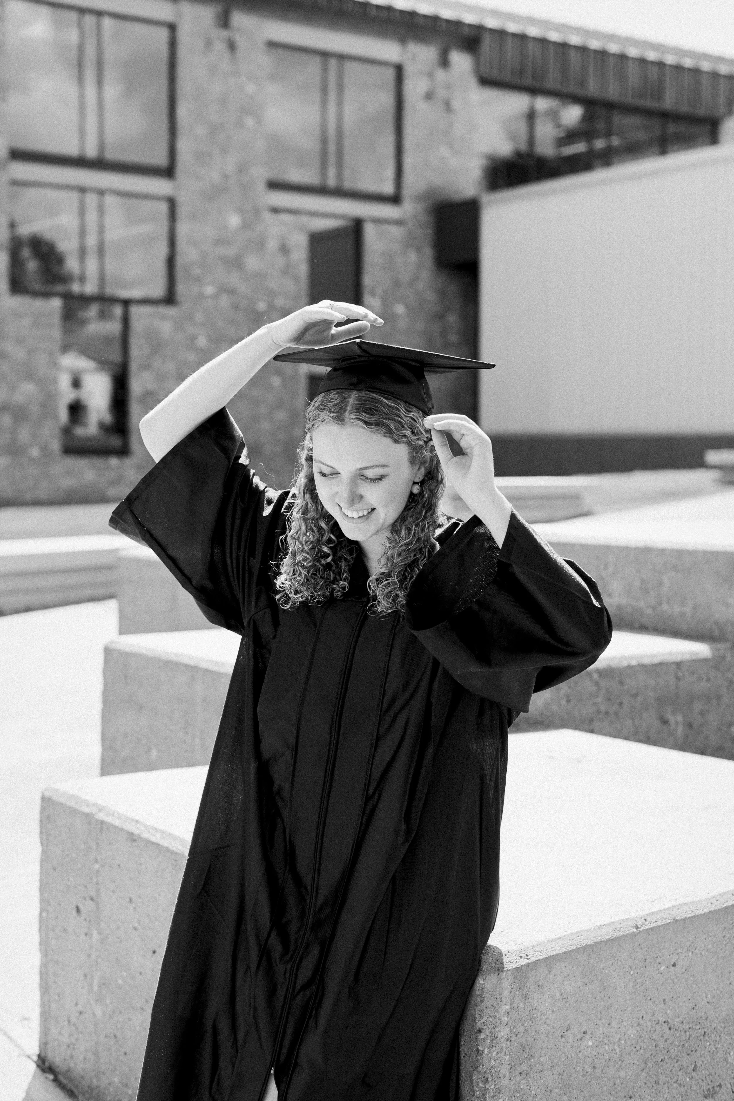 A young woman wearing a graduation gown and cap, smiling and adjusting her cap outdoors near modern buildings.