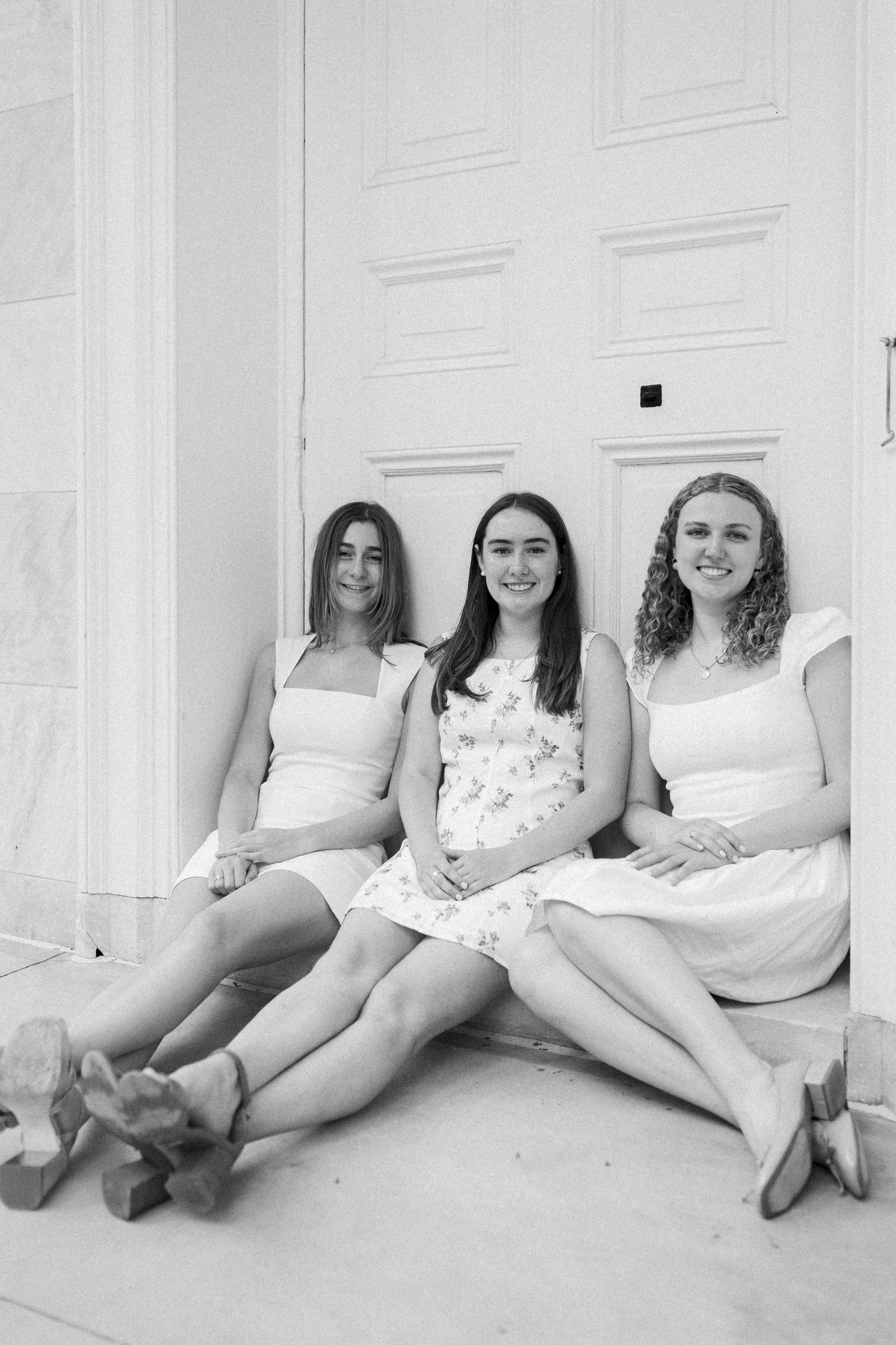 Three young women sitting on the floor against a wall with a large door behind them, smiling at the camera, in black and white.